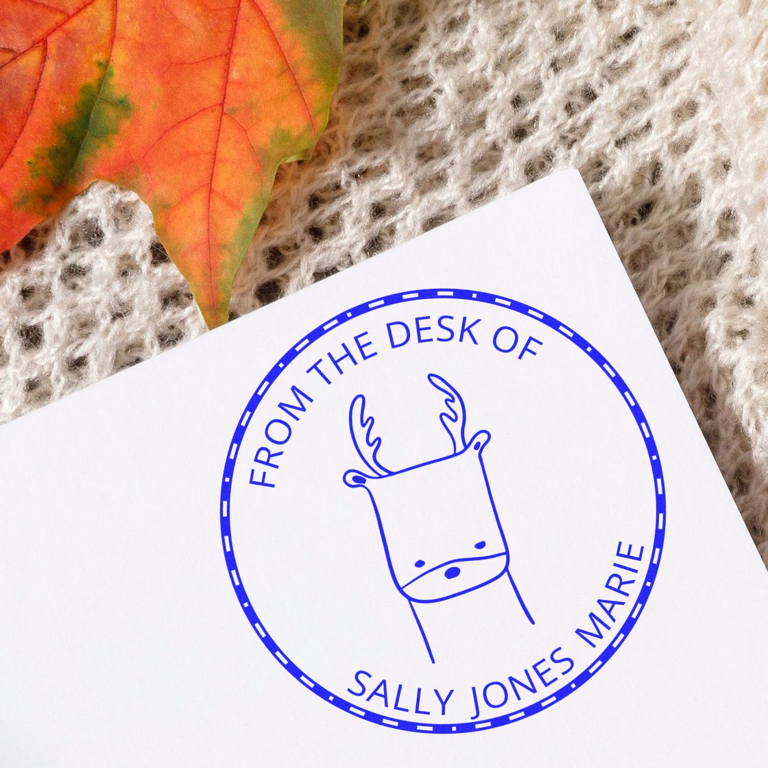 A custom-made home stamp with a wood handle, featuring a MerryMuzzle cartoon animal design, is used on a white paper. The blue circular stamp reads "From the Desk of Sally Jones Marie." A colorful autumn leaf and knitted fabric are in the background.