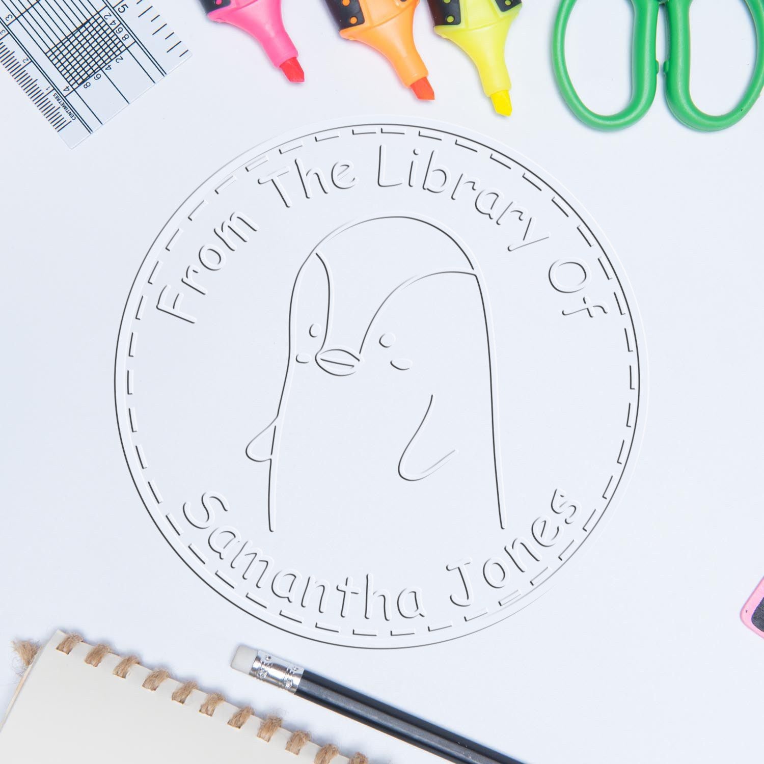 Cartoon animal desk scene featuring a PebbleWobble art embosser with a penguin design, labeled "From The Library Of Samantha Jones." Includes customizable ex libris and new address stamper options, surrounded by colorful stationery items.