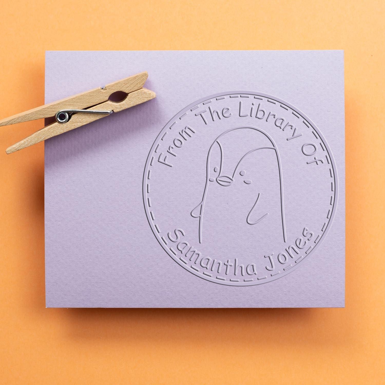 PebbleWobble Cartoon Animal Desk Personalizable Ex Libris Art Embosser featuring a cute penguin design on a purple card. Customizable New Address Stamp option available. A wooden clothespin is clipped to the card on an orange background.