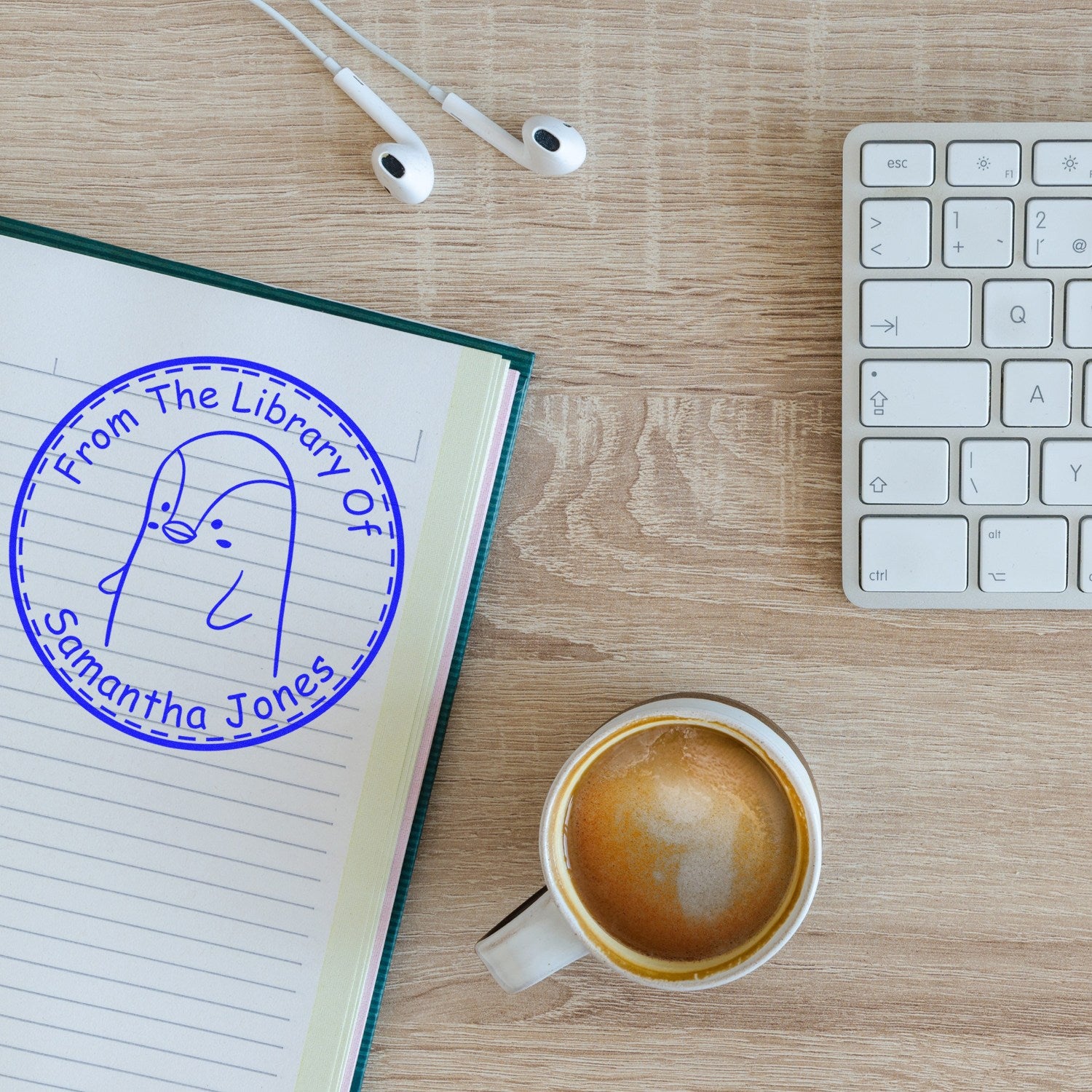 A desk with a coffee cup, keyboard, and earphones. A notebook displays a blue stamp with a cartoon animal and text "From The Library Of Samantha Jones." Keywords: Wood Handle PebbleWobble Cartoon Animal Customized Bookplate Stamp, Custom-Made New Address Stamp.
