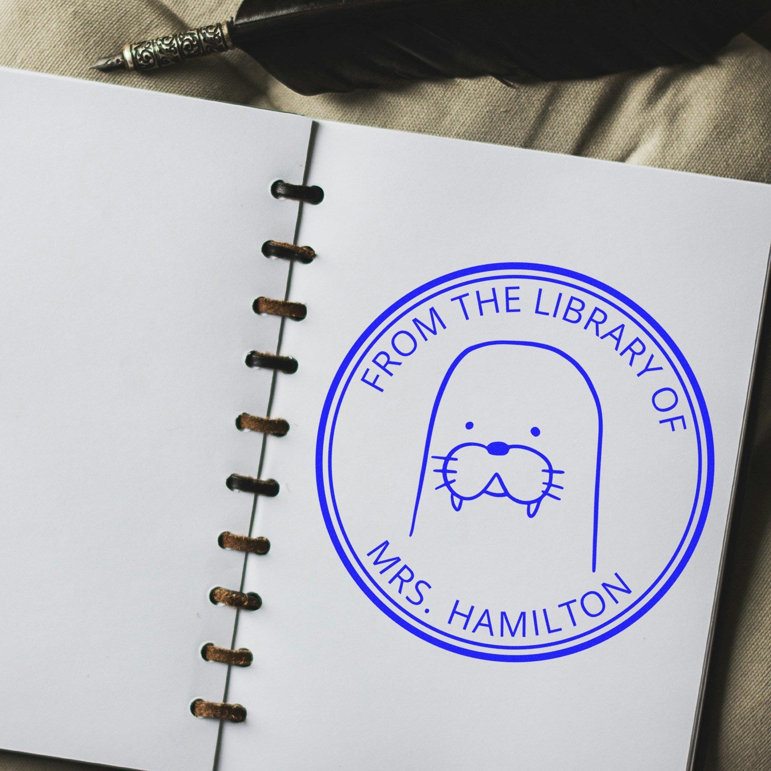 A notebook with a BumbleBristle cartoon animal pre-inked customizable Ex Libris design stamp featuring a cute seal and the text "From the Library of Mrs. Hamilton." The stamp is a custom home stamp, adding a personal touch to the page.