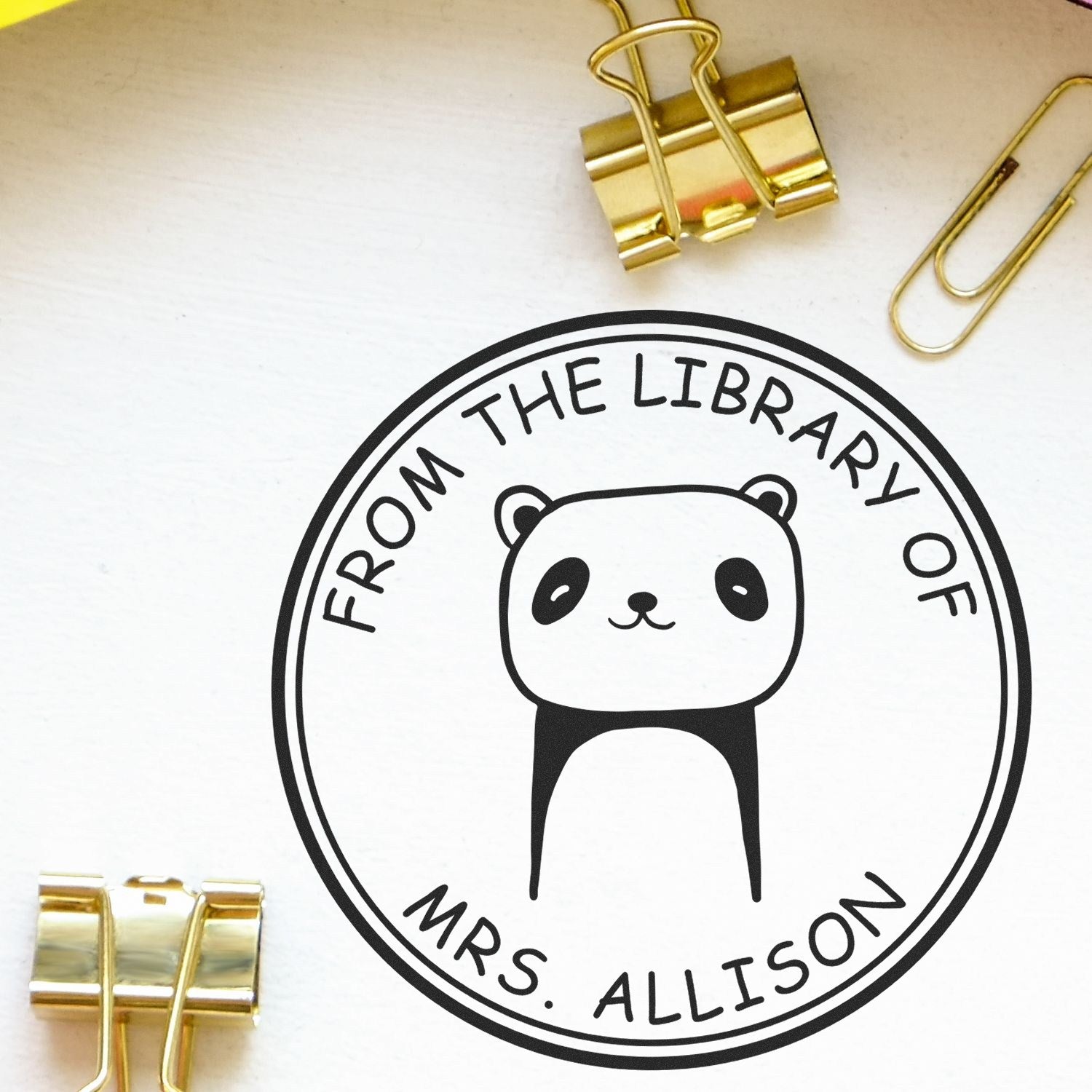 A TumbleTuft cartoon animal pre-inked customizable library bookplate stamp featuring a cute panda design with the text "From the Library of Mrs. Allison." Nearby are gold binder clips and paperclips. Ideal for custom return mail stamps.