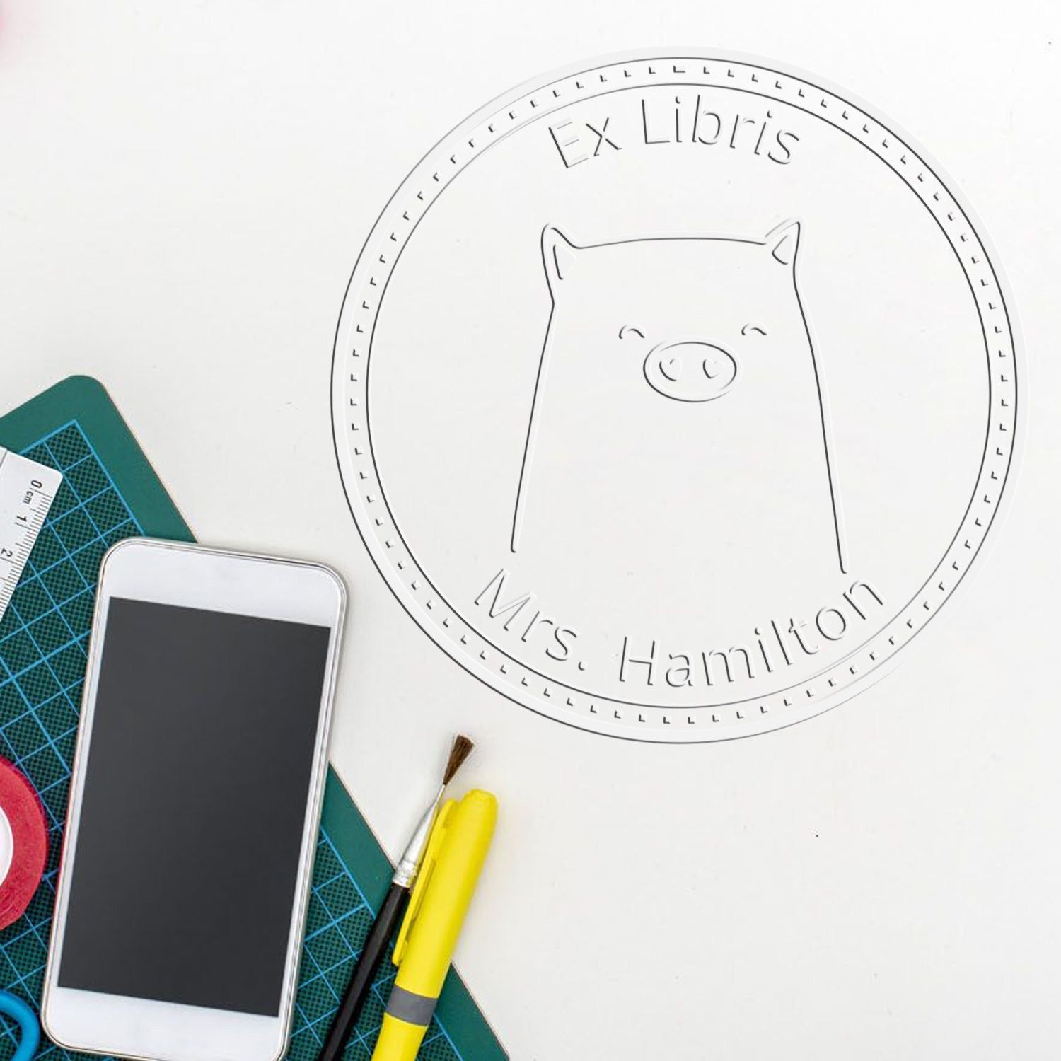 A Soft WaddleWisk cartoon animal is embossed on a custom-made book logo seal, labeled "Ex Libris Mrs. Hamilton." Nearby are a personalizable return mail stamper, a smartphone, a yellow pen, and crafting tools on a green cutting mat.