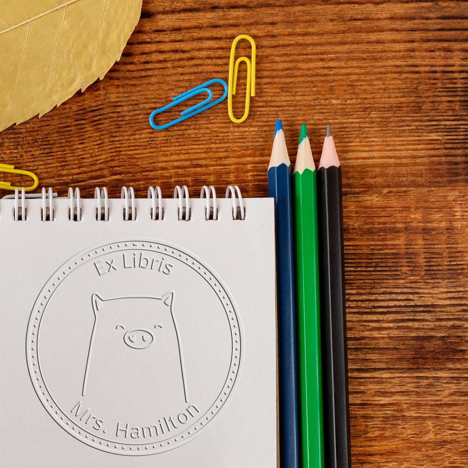 A Soft WaddleWisk cartoon animal is embossed on a custom-made book logo seal, labeled "Ex Libris Mrs. Hamilton." The personalizable mailing stamp is displayed on a notepad, surrounded by colored pencils and paperclips on a wooden surface.