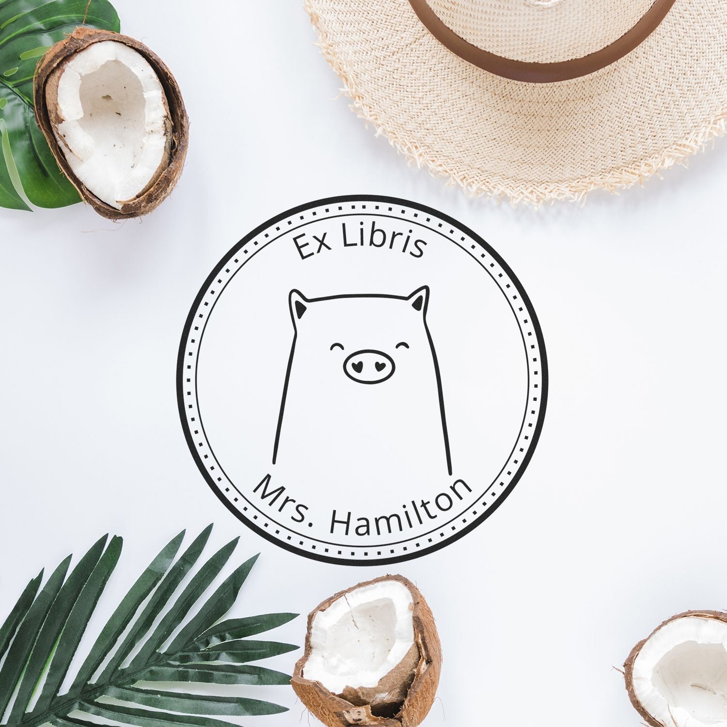A custom-made mailing stamp with a cartoon animal design, featuring a wood handle WaddleWisk. The stamp reads "Ex Libris Mrs. Hamilton" and is placed on a white surface surrounded by tropical leaves, coconut shells, and a straw hat.