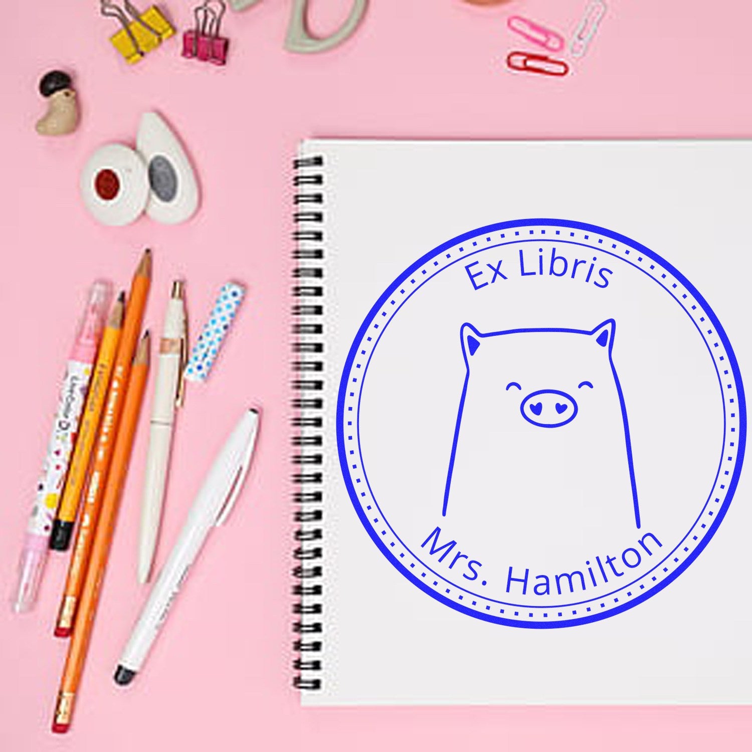 A pink desk with stationery items surrounds a spiral notebook featuring a blue cartoon animal stamp labeled Ex Libris Mrs. Hamilton. The stamp, a custom-made mailing stamper, has a wood handle WaddleWisk design customized from the desk of stamp.