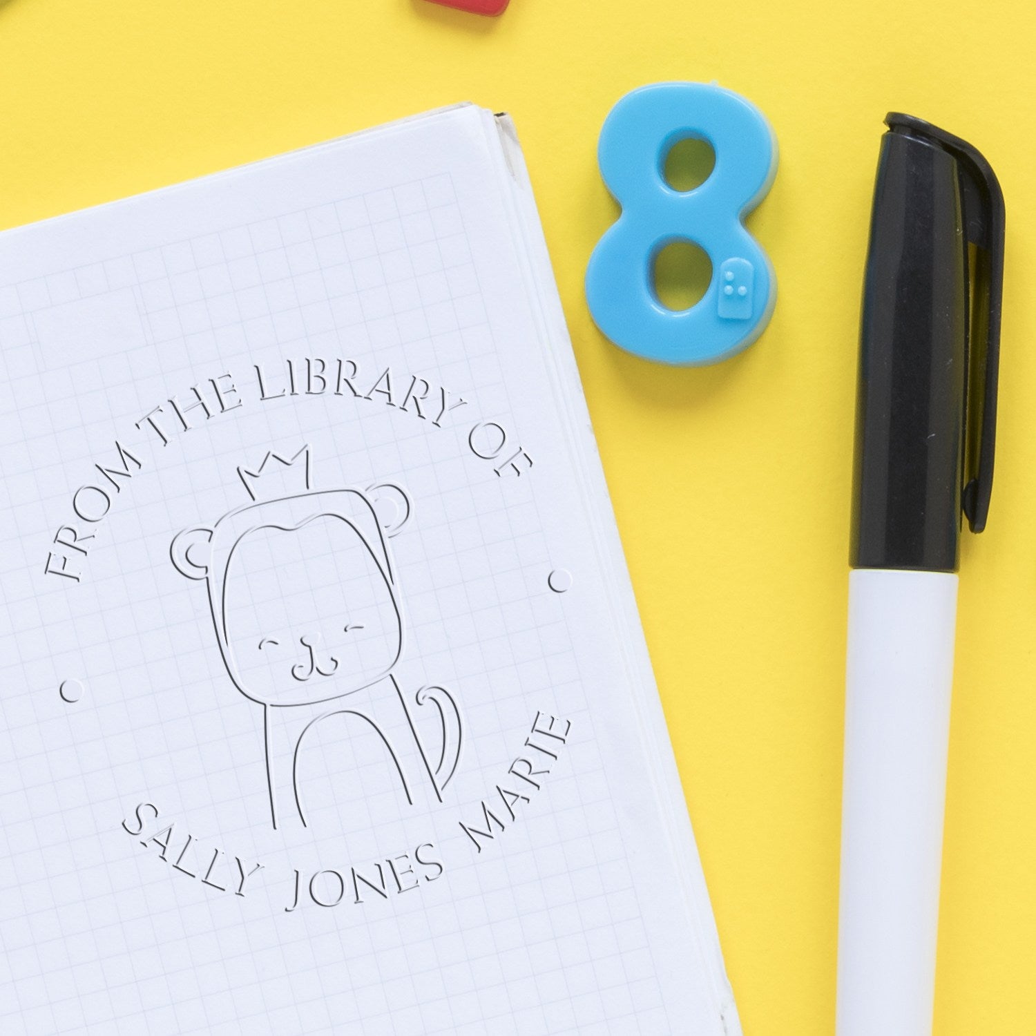 A ScribbleScrabble cartoon animal hybrid embossing seal is stamped on a notebook page, reading "From the Library of Sally Jones Marie." Nearby are a blue number eight and a black pen. Ideal for a personalized new address stamper.