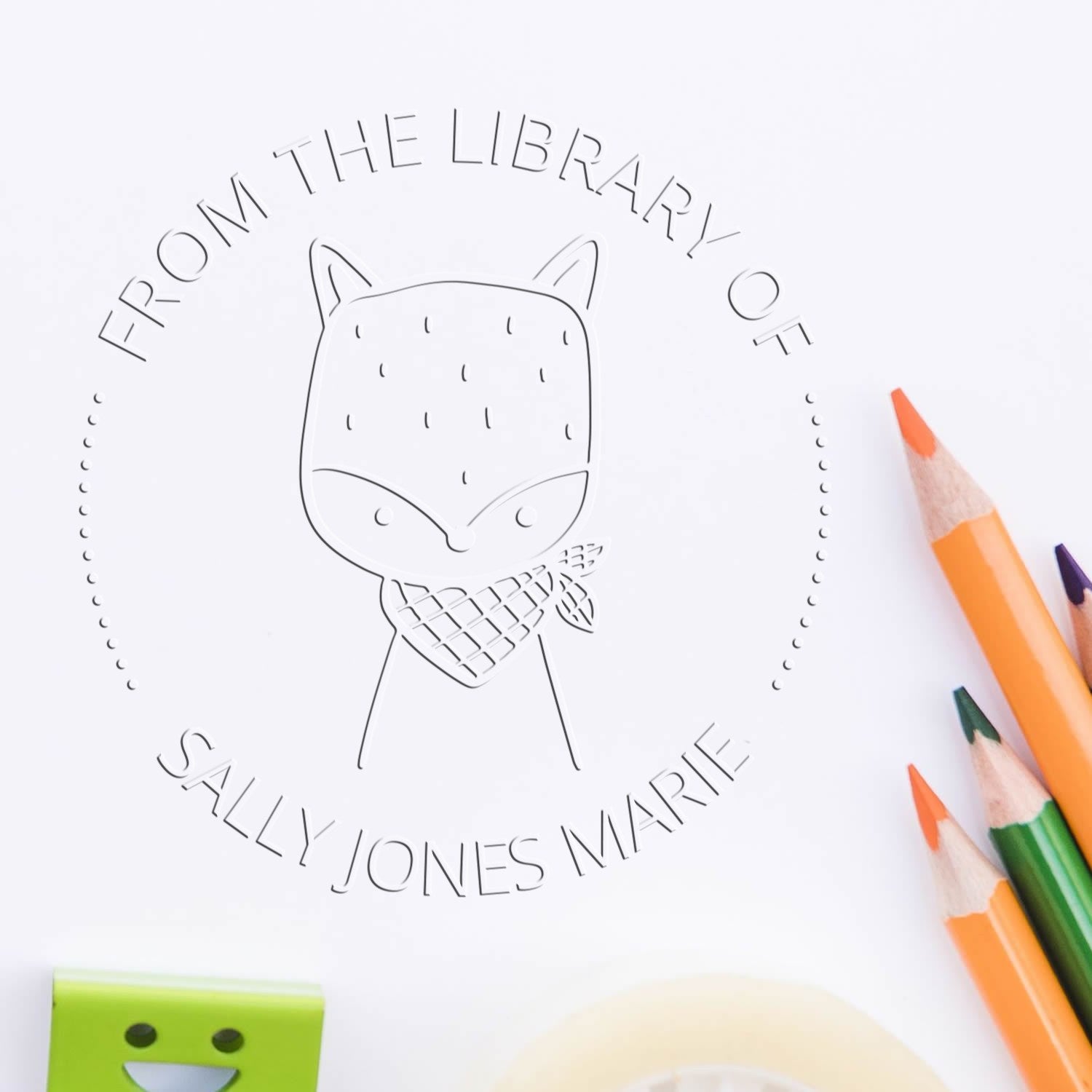 A custom-made book insignia seal featuring a Soft SnickerSnack cartoon animal with a scarf, labeled "From the Library of Sally Jones Marie." Colored pencils and a customizable return mail stamper are nearby on a white surface.
