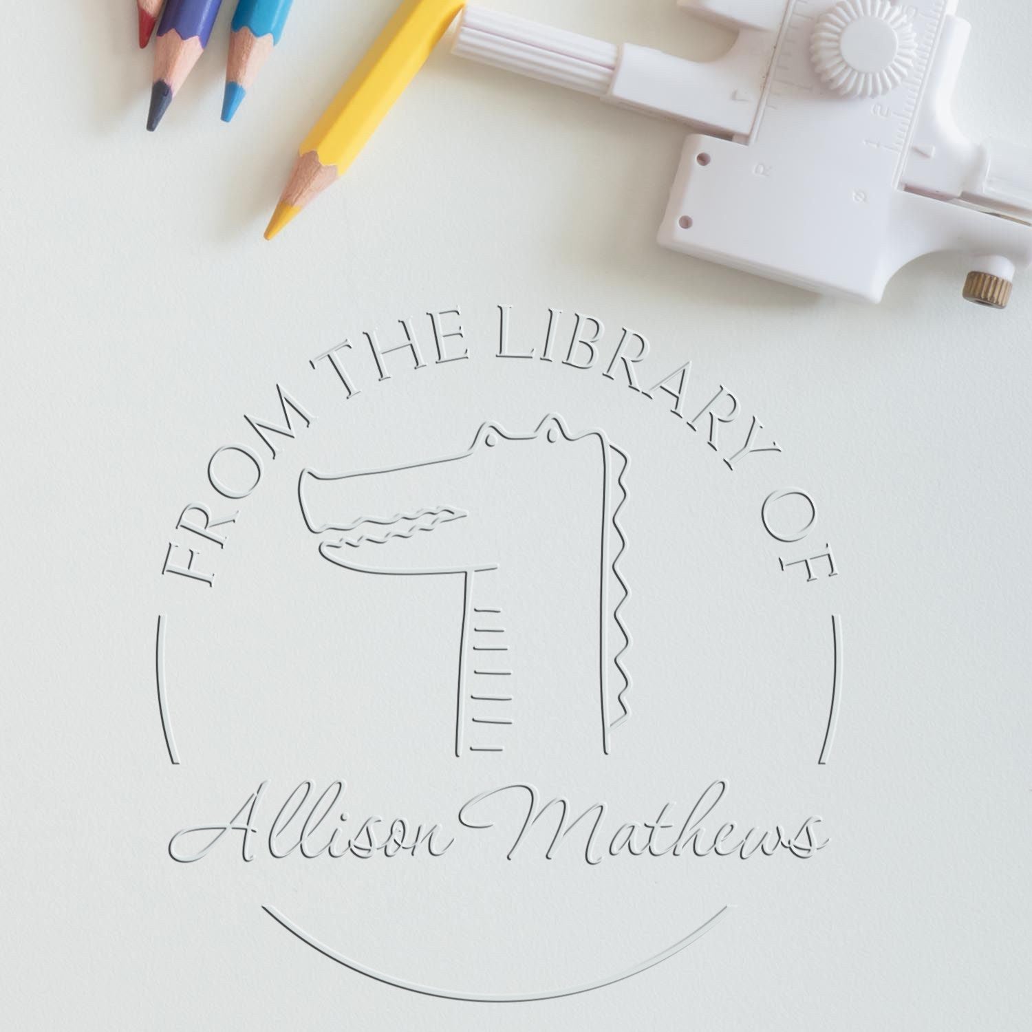 NoodleNose cartoon animal design embossed on paper using a handheld personalized home library embosser. Colored pencils are nearby. The embosser is also a personalizable mailing stamper.