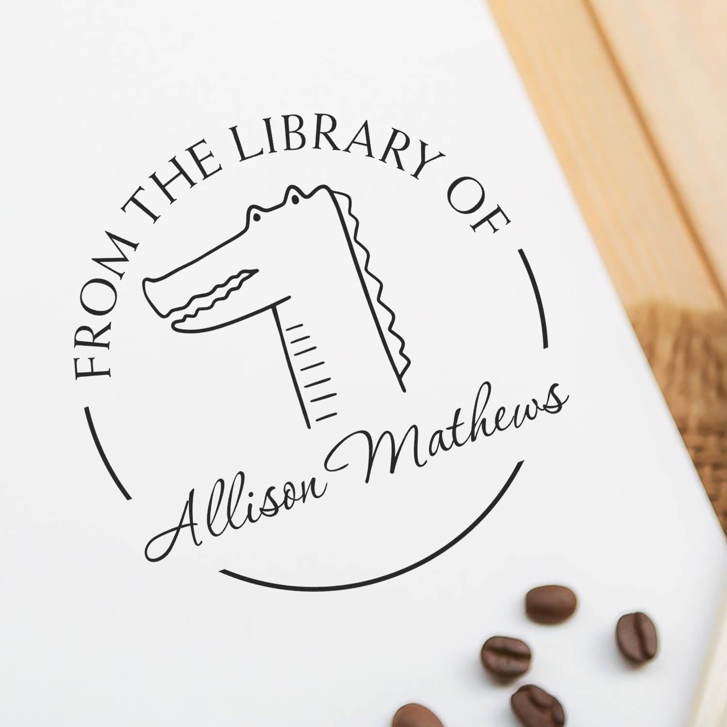 NoodleNose Cartoon Animal Pre-Inked Customizable Ex Libris Book Stamp featuring a stylized animal design with the text "From the Library of Allison Mathews." Nearby are coffee beans on a wooden surface. Ideal for a Customized Address Stamper.
