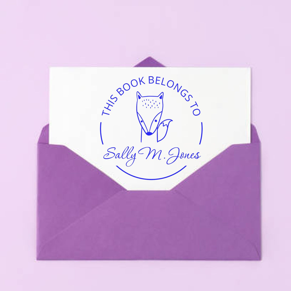 A purple envelope contains a card with a Slim Pre-Inked BlinkBounce Cartoon Animal Custom Library Book With Name Stamp design, featuring a fox and the text "This Book Belongs To Sally M. Jones." Ideal for a Customizable Return Mail Stamp.