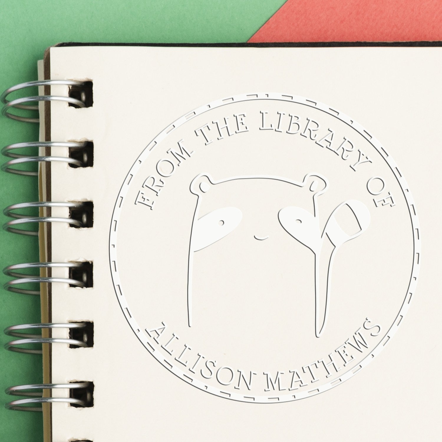 A personalized return mail stamper featuring a Soft FreckleFlop cartoon animal design. The custom-made book branding seal is embossed on a notebook page, with the text From the Library of Allison Mathews.
