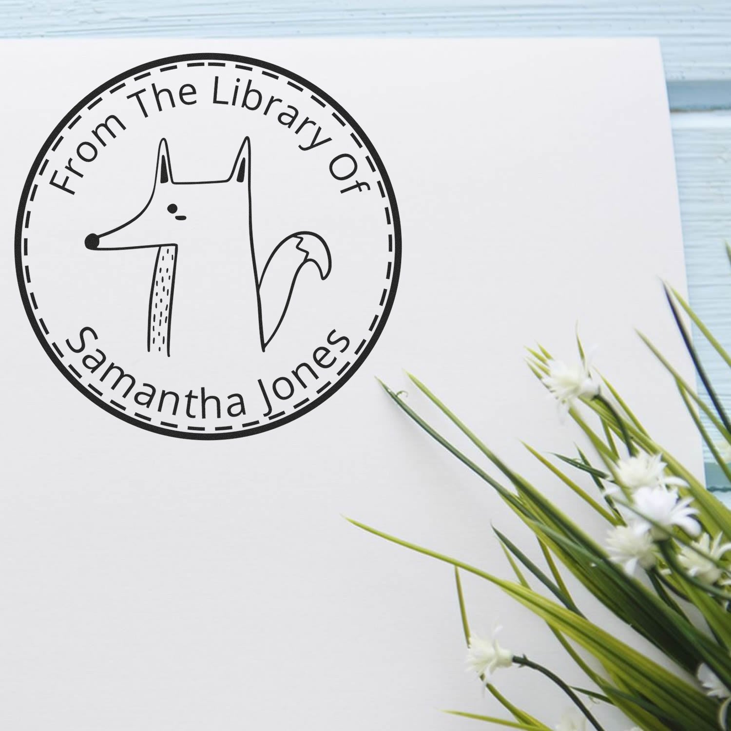 Image of a Wood Handle CuddleCub Cartoon Animal Customized Book Stamp on paper, featuring a fox illustration with text "From The Library Of Samantha Jones." Nearby, a plant is visible. This stamp is also a Customizable New Address Stamp.