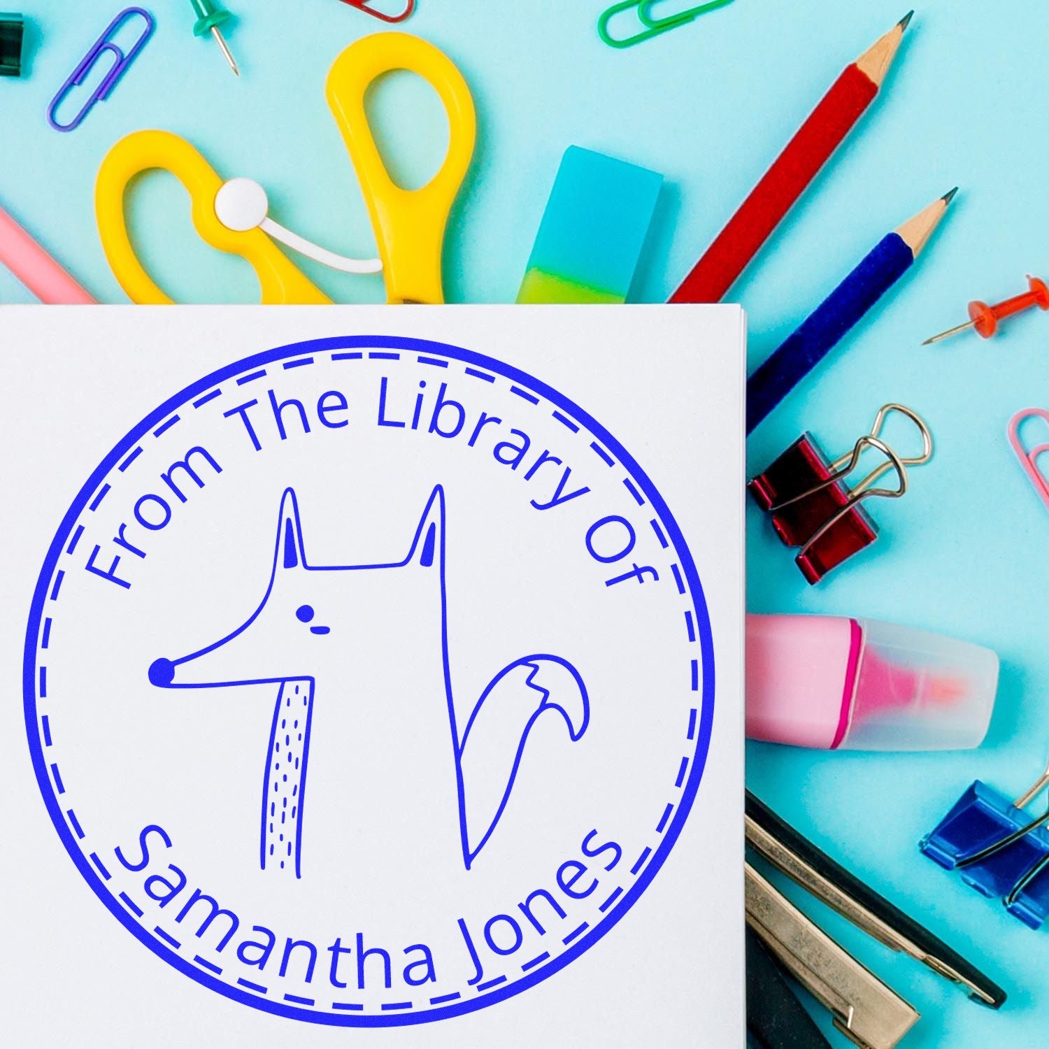 Slim Pre-Inked CuddleCub Cartoon Animal Custom Book Ownership Stamp displayed on a white paper, surrounded by colorful stationery. The custom stamp features a cute fox illustration with the text "From The Library Of Samantha Jones" in blue.