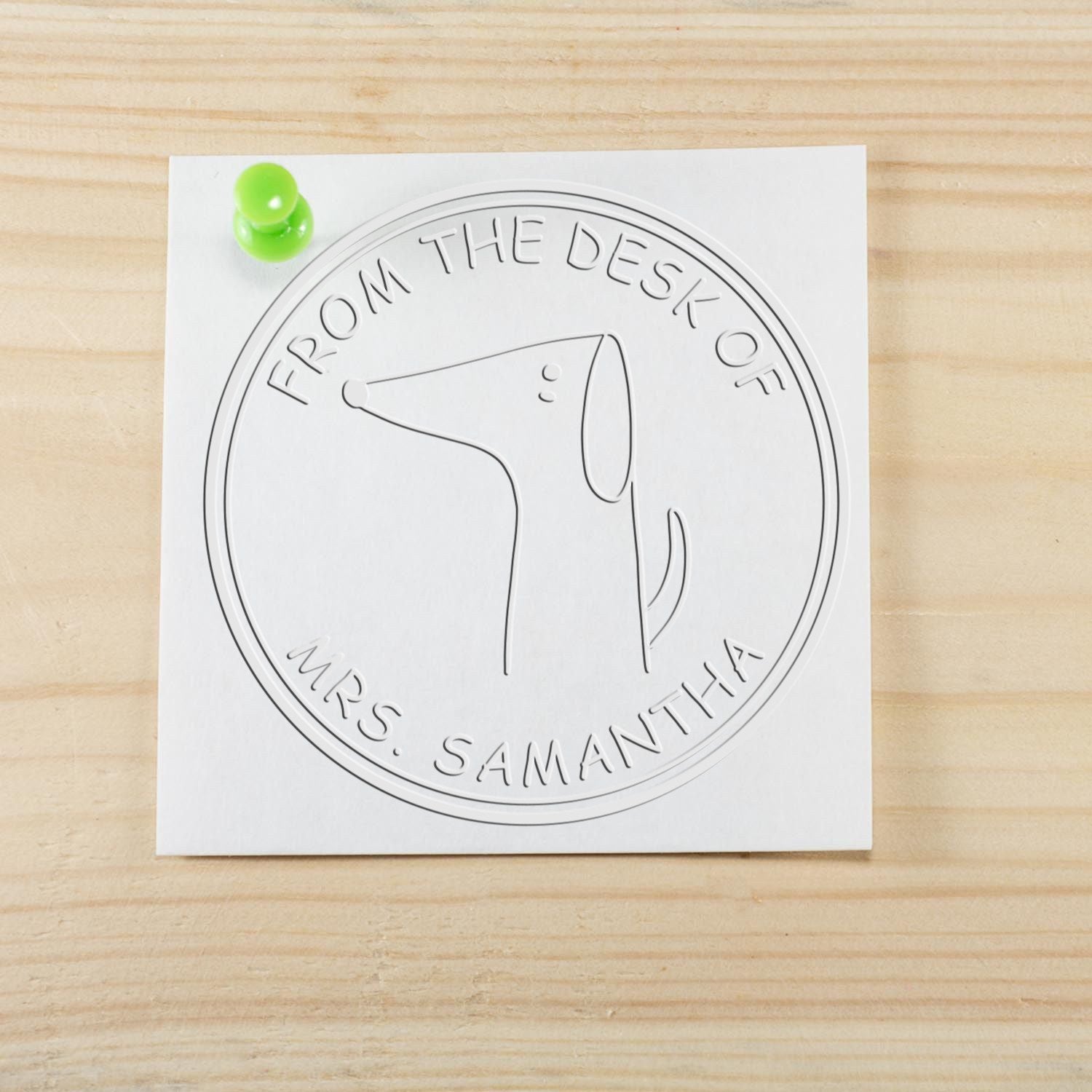 Embossed paper with a Soft WiggleWag cartoon animal design, featuring the text "From the Desk of Mrs. Samantha." The custom-made book identity embosser and personalized address stamp are displayed on a wooden surface with a green pushpin.