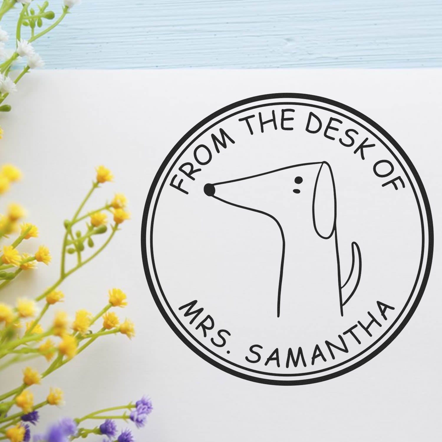 Image of a customizable new address stamper featuring a wood handle WiggleWag cartoon animal design. The stamp, used on white paper, reads "From the Desk of Mrs. Samantha" and is surrounded by small yellow and purple flowers.