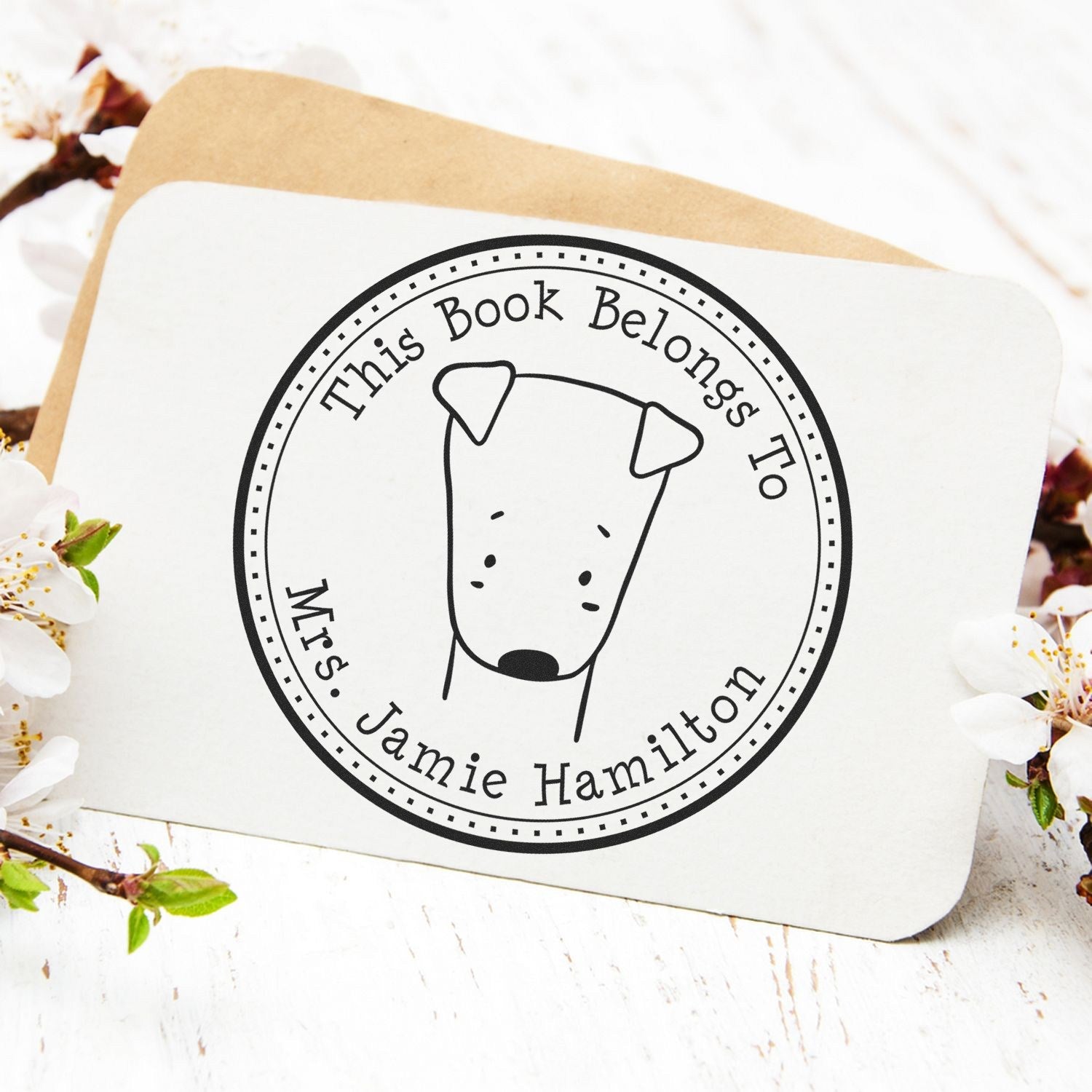 Wood handle SizzleSniff cartoon animal customized book monogram stamp on a card, featuring a simple animal illustration. The custom mailing stamper is surrounded by blooming flowers on a light wooden surface.