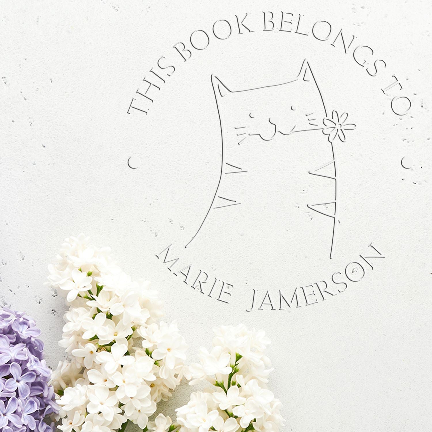 Soft HuddleHopper cartoon animal design on a custom-made book personalization embosser. The embosser features a cat with a flower, surrounded by text. Nearby are lilac and white flowers. Includes a custom return mail stamp.
