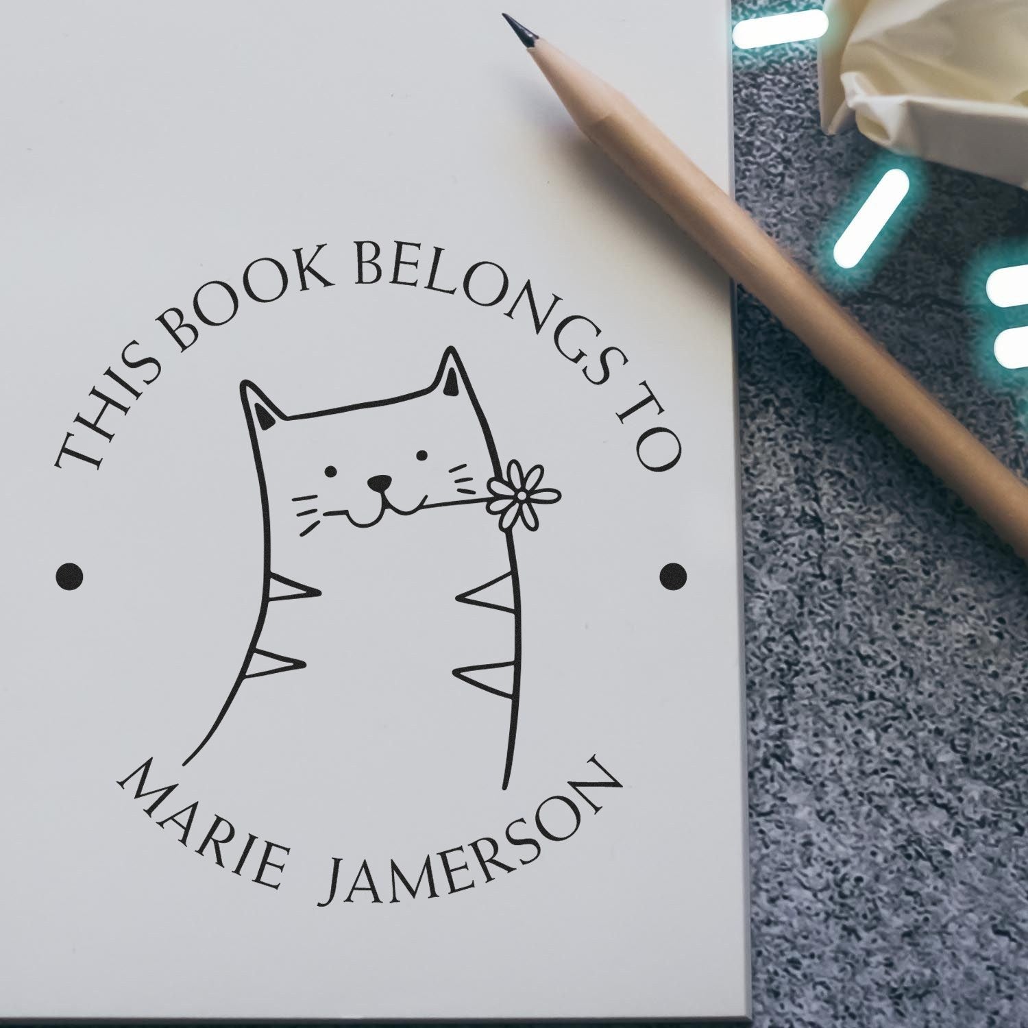 HuddleHopper Cartoon Animal Pre-Inked Customizable Book Mark Stamp featuring a cute cat design with a flower, labeled "This Book Belongs To Marie Jamerson." The Customized Home Stamp is shown on a notebook with a pencil nearby.
