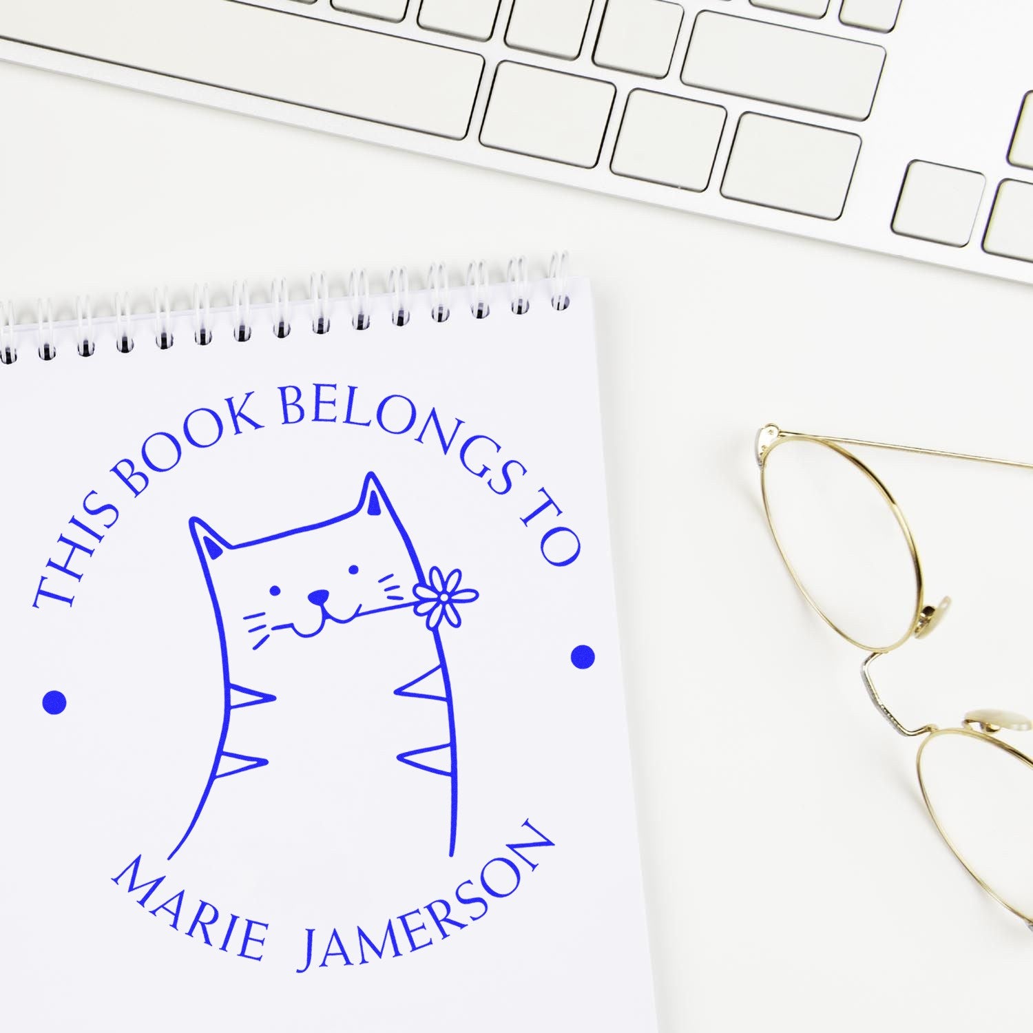 HuddleHopper Cartoon Animal Pre-Inked Customizable Book Mark Stamp featuring a cute cat design on a notebook. Beside it, a Customized Home Stamper is placed near a keyboard and glasses.