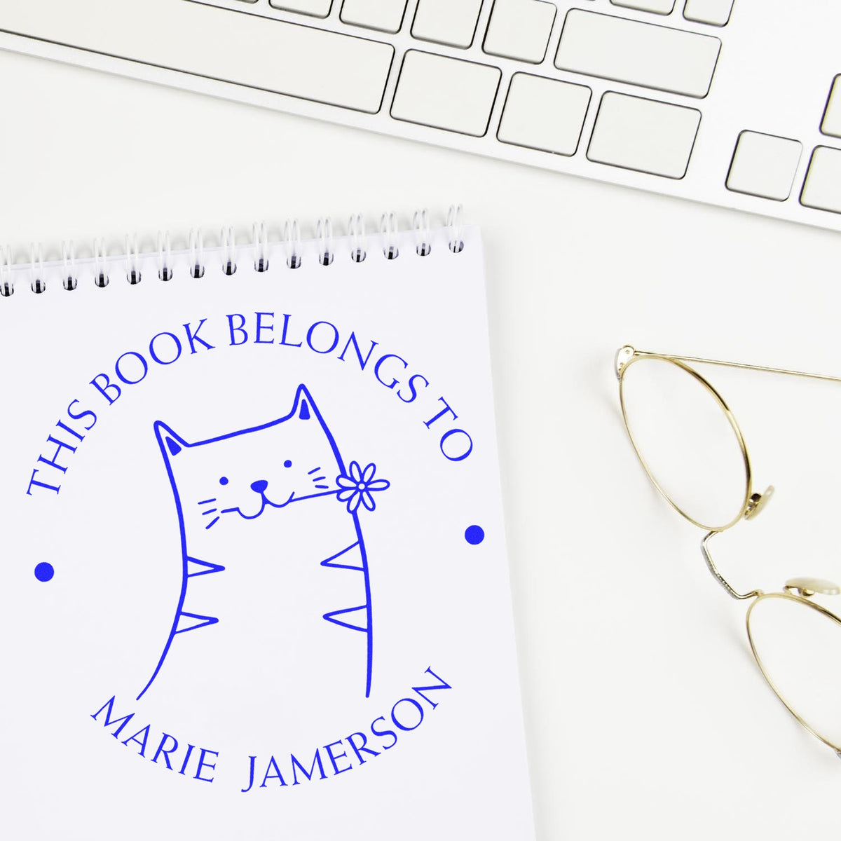 Slim Pre-Inked HuddleHopper Cartoon Animal Custom from the Study of Stamp displayed on a notebook page. The customizable mailing stamp features a cute cat holding a flower, with text "This Book Belongs To Marie Jamerson" in blue ink.