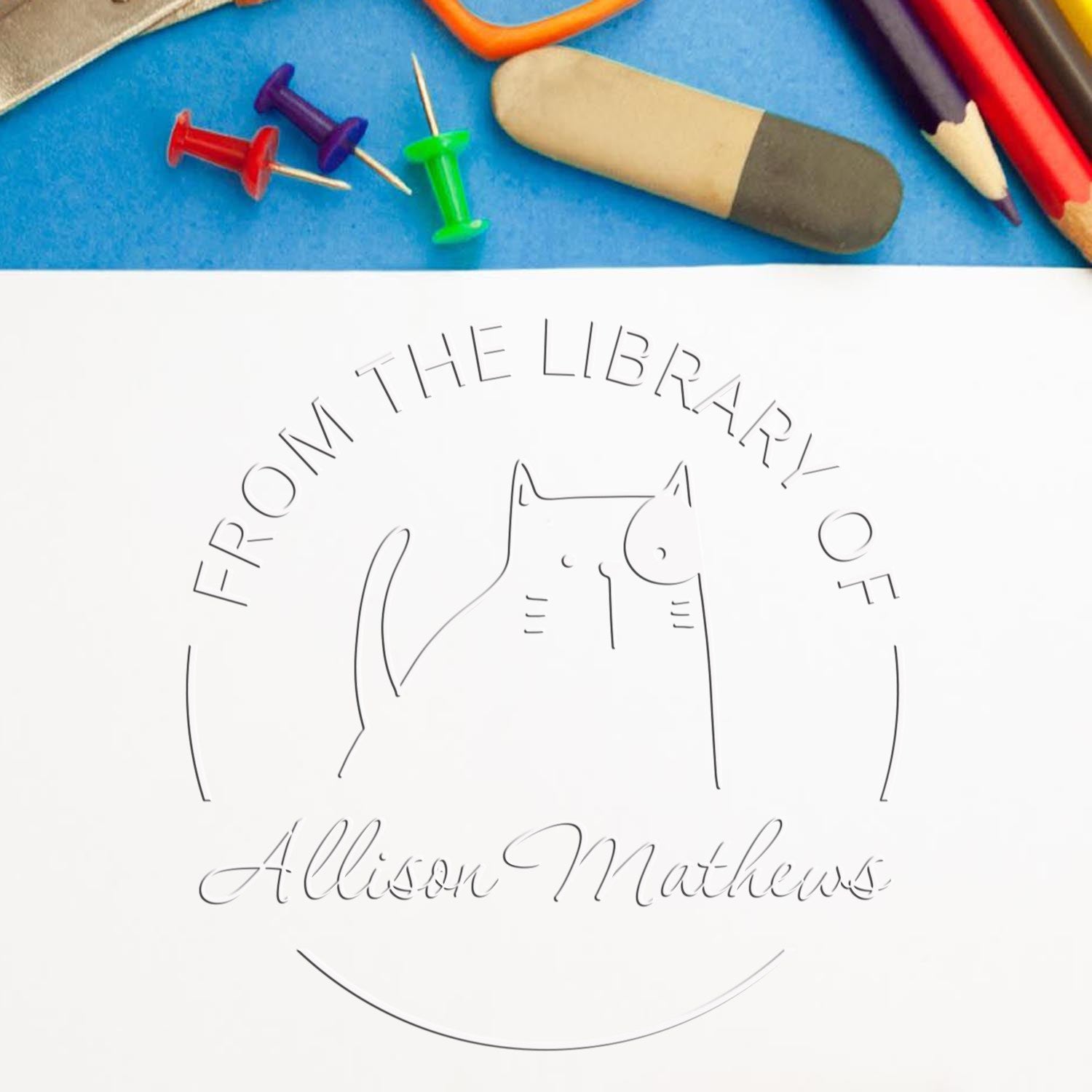 A GobbleGiggle cartoon animal design is embossed on a white paper using a handheld personalized book mark embosser. Nearby are colorful push pins, pencils, and an eraser. The text reads "From the Library of Allison Mathews." Personalized address stamper not shown.