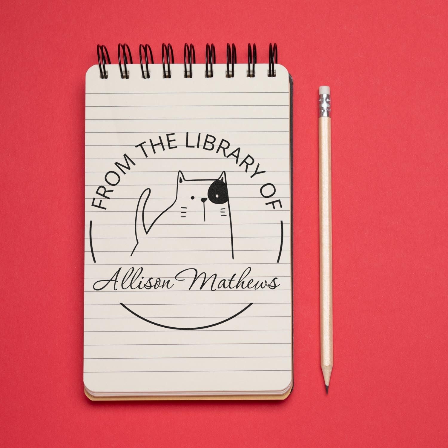 A notebook with a GobbleGiggle Cartoon Animal Pre-Inked Customizable Library Book Belongs To Stamp featuring a cat design and the name "Allison Mathews." A pencil lies beside it. Ideal for a Personalizable Address Stamp.