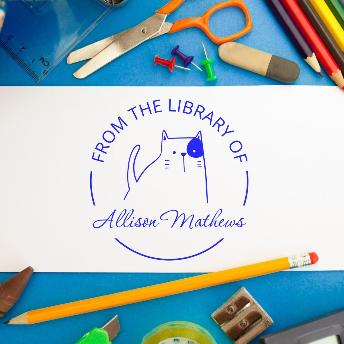 A Slim Pre-Inked GobbleGiggle Cartoon Animal Custom Library Bookplate Stamp is shown on a white paper, featuring a cute cat design with "From the Library of" text. Surrounding the paper are colorful office supplies. Ideal as a Personalized Home Stamp.