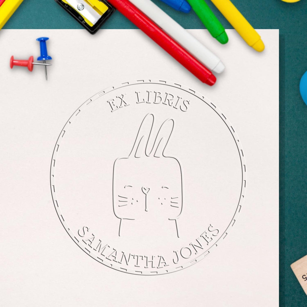 Cartoon animal hybrid "SquirmSquiggle" embossed on a library book page with the name "Samantha Jones" in a seal. Surrounding items include colorful pens and a customized mailing stamper.