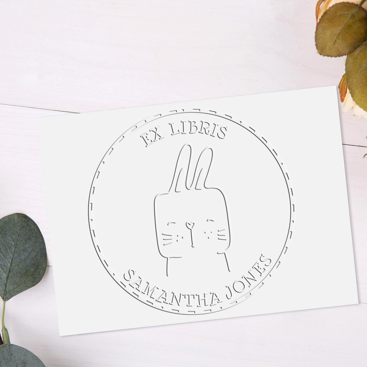 A SquirmSquiggle cartoon animal hybrid is embossed on a library book with a name embossing seal reading "Ex Libris Samantha Jones." The design resembles a customized mailing stamp, featuring a cute animal with long ears.