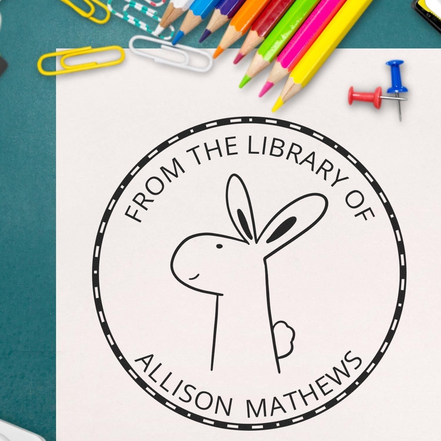 Cartoon animal stamp with a rabbit design labeled "From the Library of Allison Mathews" on paper. Surrounded by colorful pencils and paperclips. Keywords: BlinkerBelle Cartoon Animal Self-Inking Personalizable This Book Belongs To Stamp, Custom New Address Stamp.