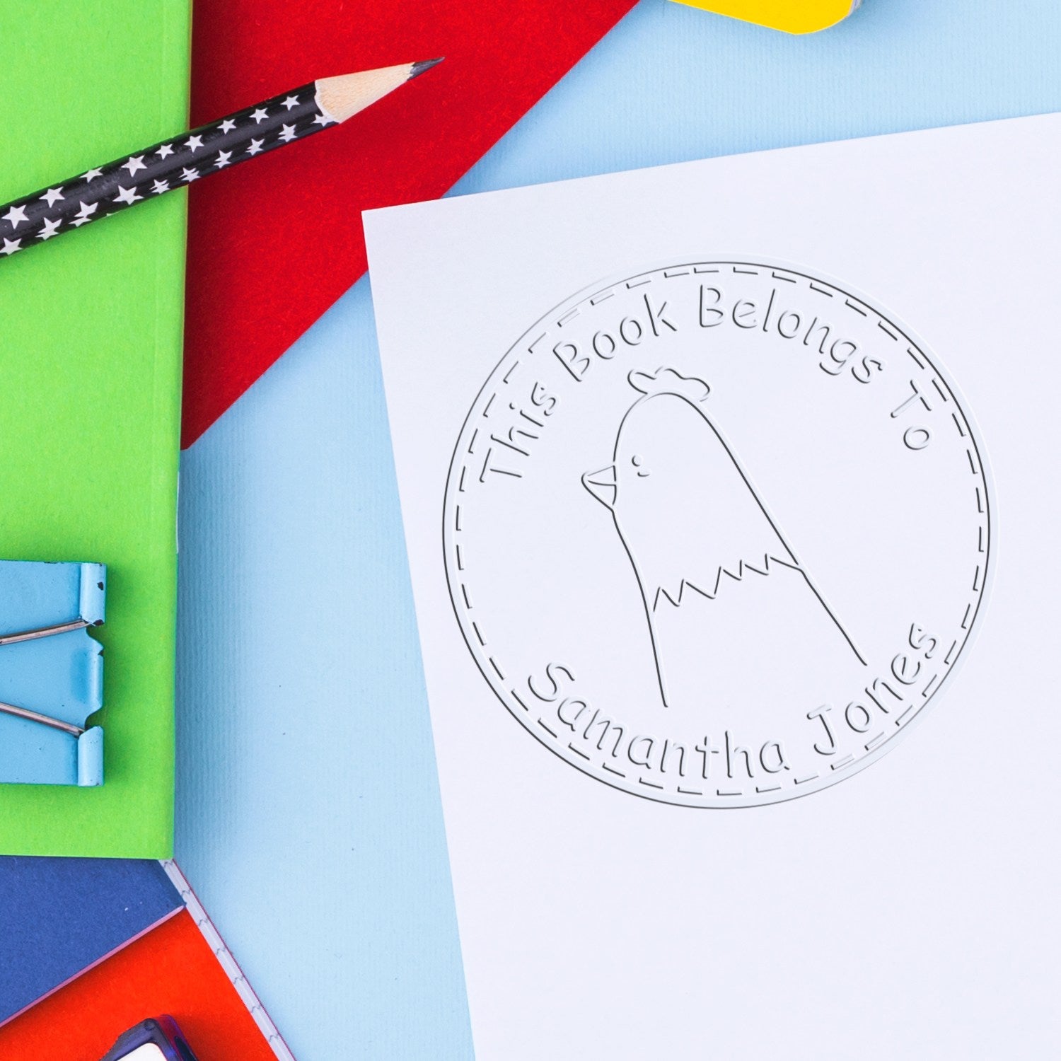 A FuzzleFeather cartoon animal embosser is shown on a white paper, featuring a chicken design with the text "This Book Belongs To Samantha Jones." Nearby are colorful folders and clips. Keywords: Personalized Mailing Stamper, Handheld Personalized Book Belongs To Embosser.