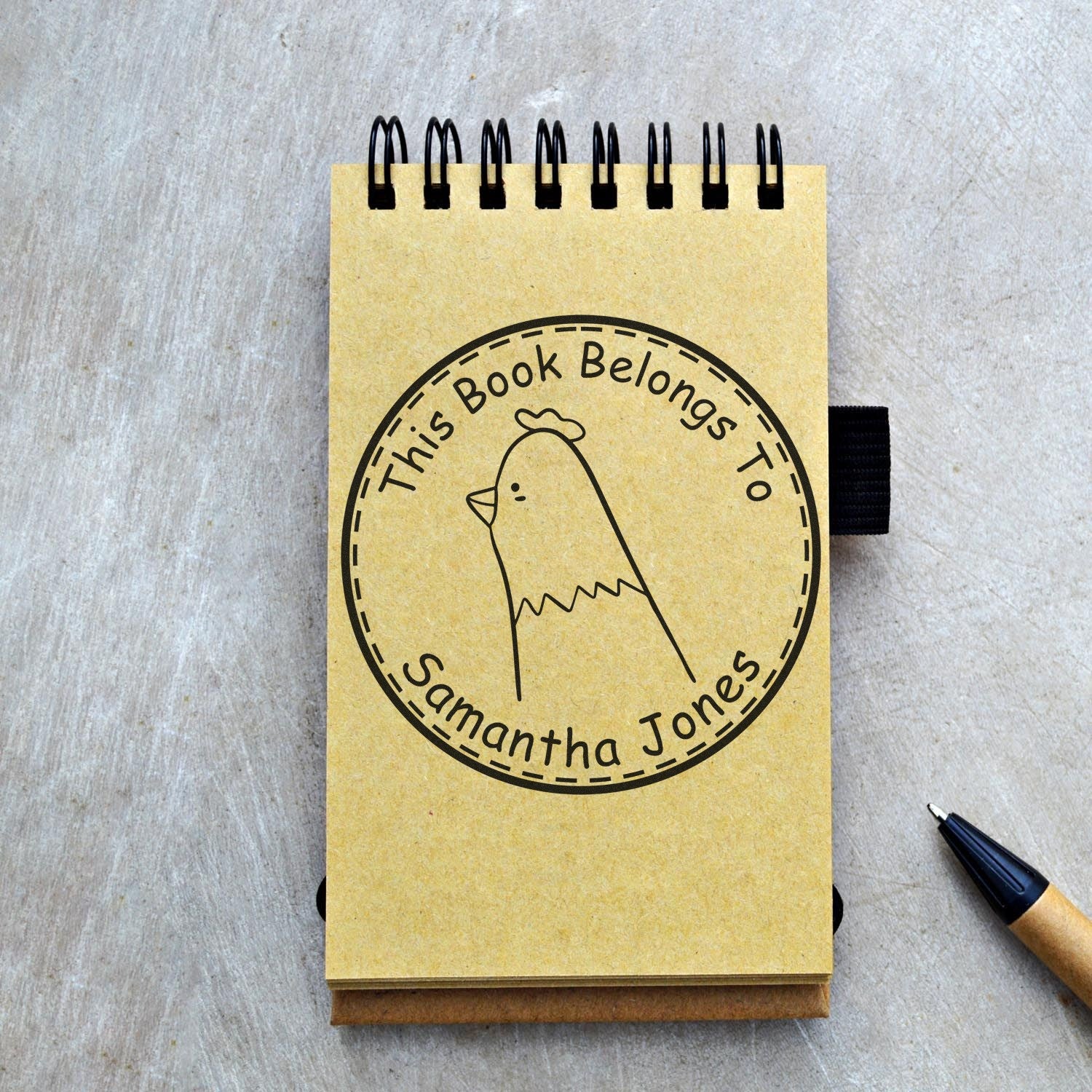 A notebook with a FuzzleFeather cartoon animal pre-inked customizable stamp on the cover, reading "This Book Belongs To Samantha Jones." The custom mailing stamp design features a simple bird illustration. A pen is placed beside the notebook.