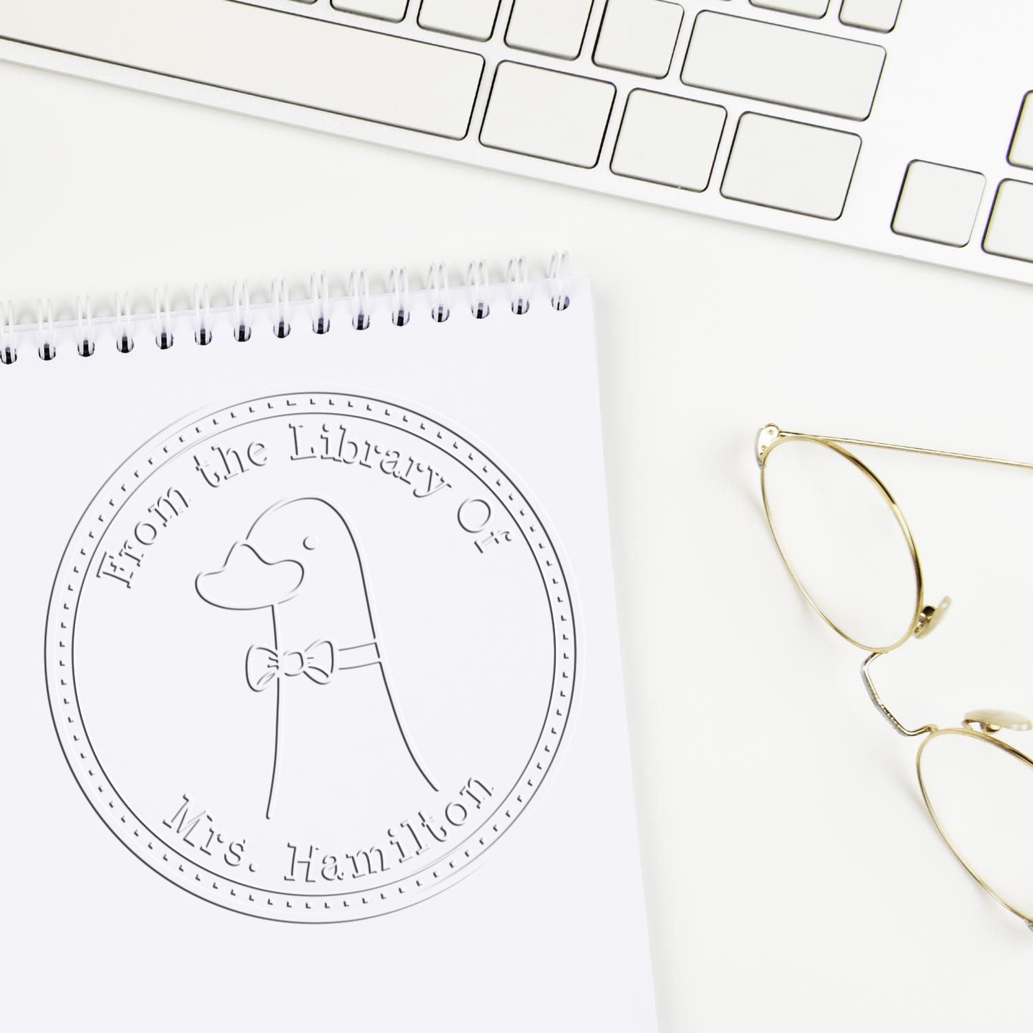 A DoodleDuck cartoon animal embossing seal is pressed onto a notebook page, displaying "From the Library Of Mrs. Hamilton." Nearby are a keyboard and glasses. Keywords: DoodleDuck Cartoon Animal Handheld Personalized Book Belongs To Embossing Seal, Custom-Made Mailing Stamper.