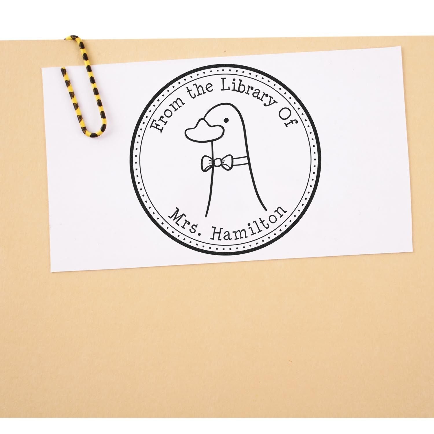 DoodleDuck cartoon animal self-inking personalizable book return stamp on a card, featuring a duck with a bow tie. The text reads "From the Library Of Mrs. Hamilton." Custom-made new address stamp design on a beige background with a black and yellow string.