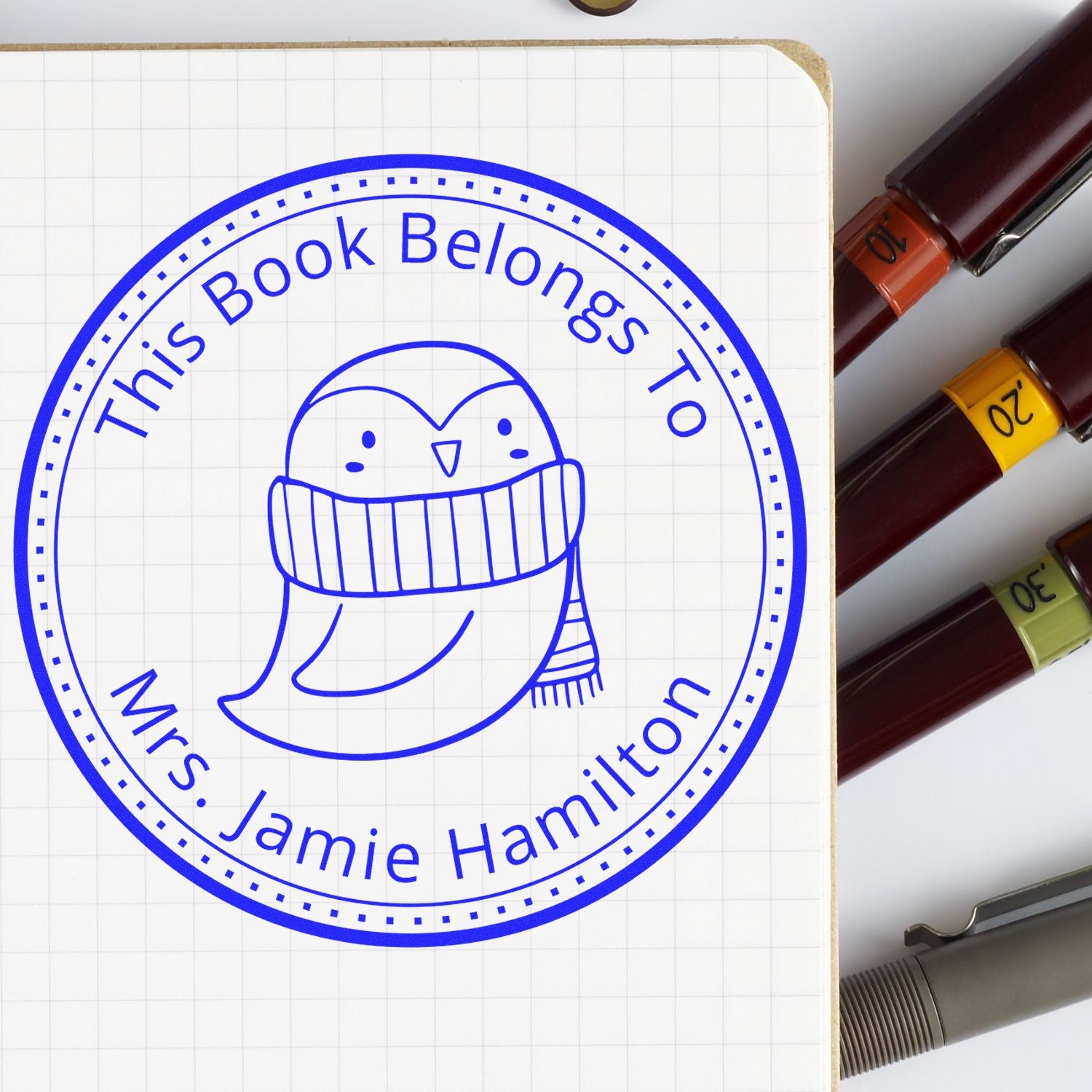 Wood handle stamp featuring a QuirkyQuack cartoon animal design on a customized bookplate. The stamp reads 'This Book Belongs To Mrs. Jamie Hamilton.' Includes customizable mailing stamp options, shown next to pens on a grid notebook.