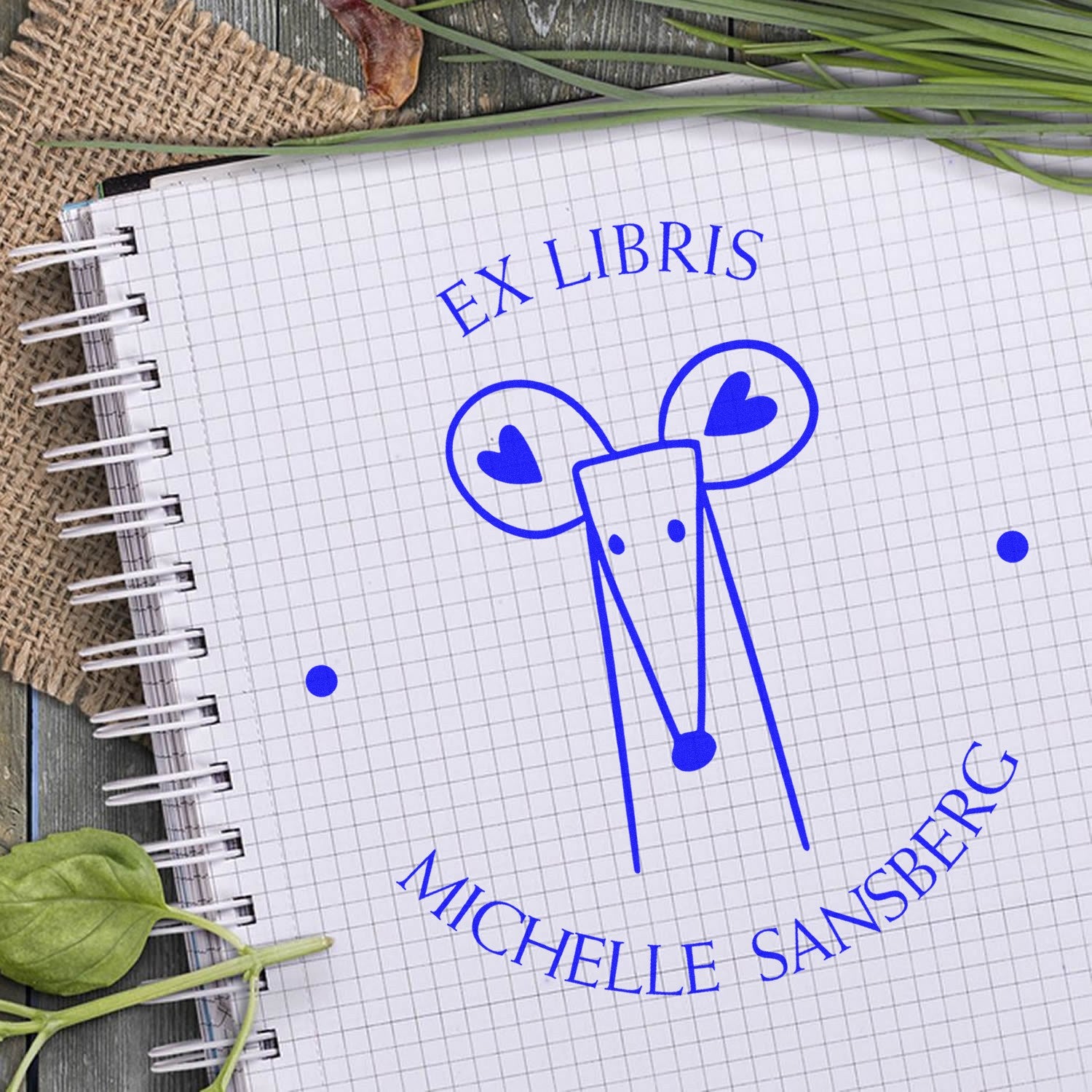Slim Pre-Inked DazzleDash Cartoon Animal Custom From The Office Of Stamp on a notebook. The design features a stylized animal with hearts, surrounded by text. Ideal for a Customizable Return Mail Stamp. Notebook on a wooden surface with leaves.