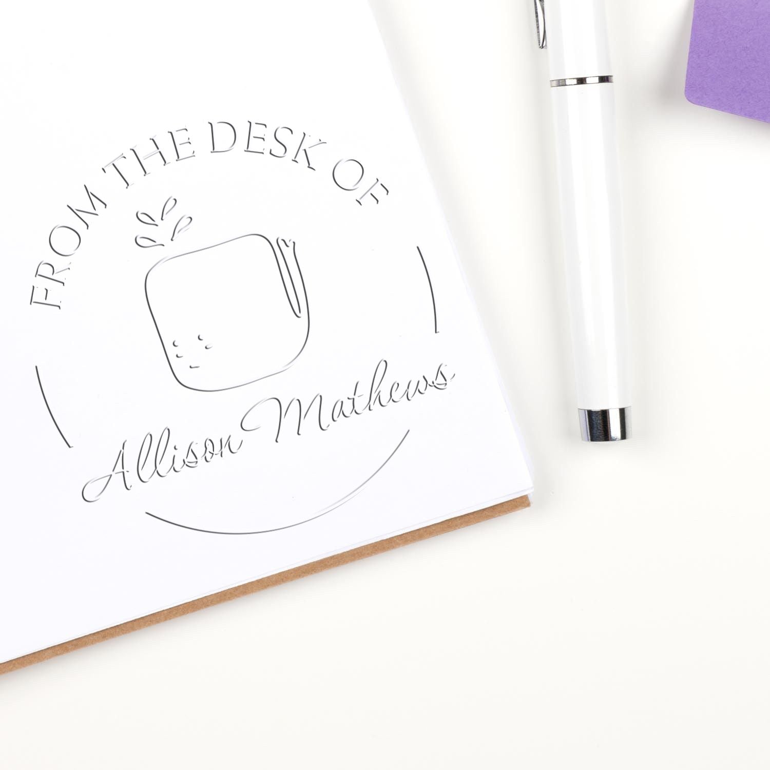 A WhiffleWhirl cartoon animal design is embossed on a paper using a handheld personalized library embosser. The text reads "From the Desk of Allison Mathews." A pen and a purple notepad are nearby. Custom return mail stamper is also visible.