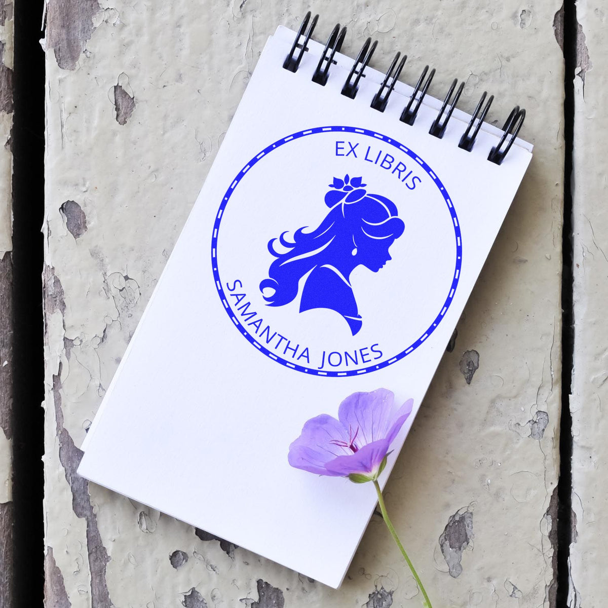 White spiral notebook featuring a blue ex libris seal stamp and purple flower on distressed gray wood.