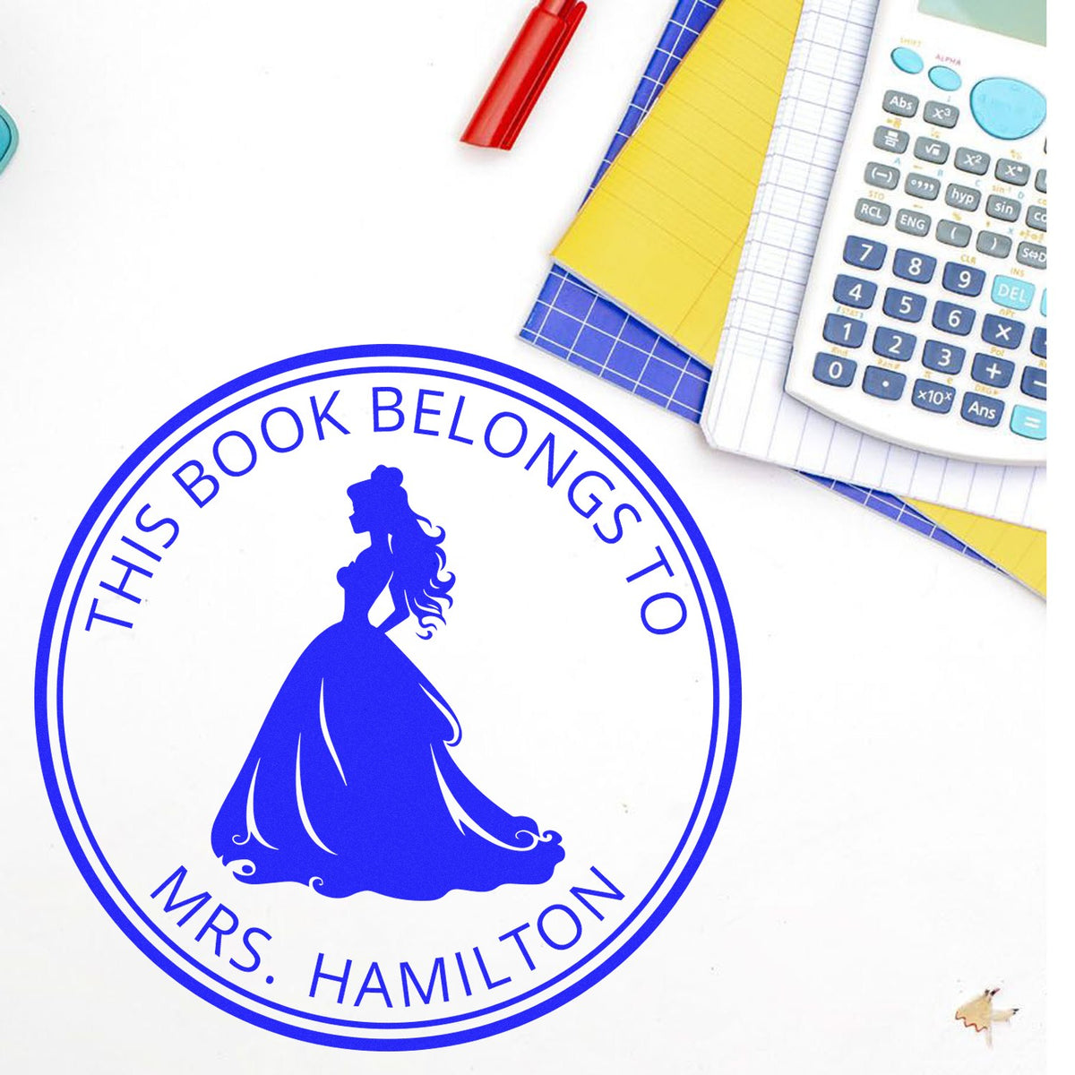 Blue custom book-belongs-to stamp seal with princess silhouette, beside a white calculator, notebooks, and red pen