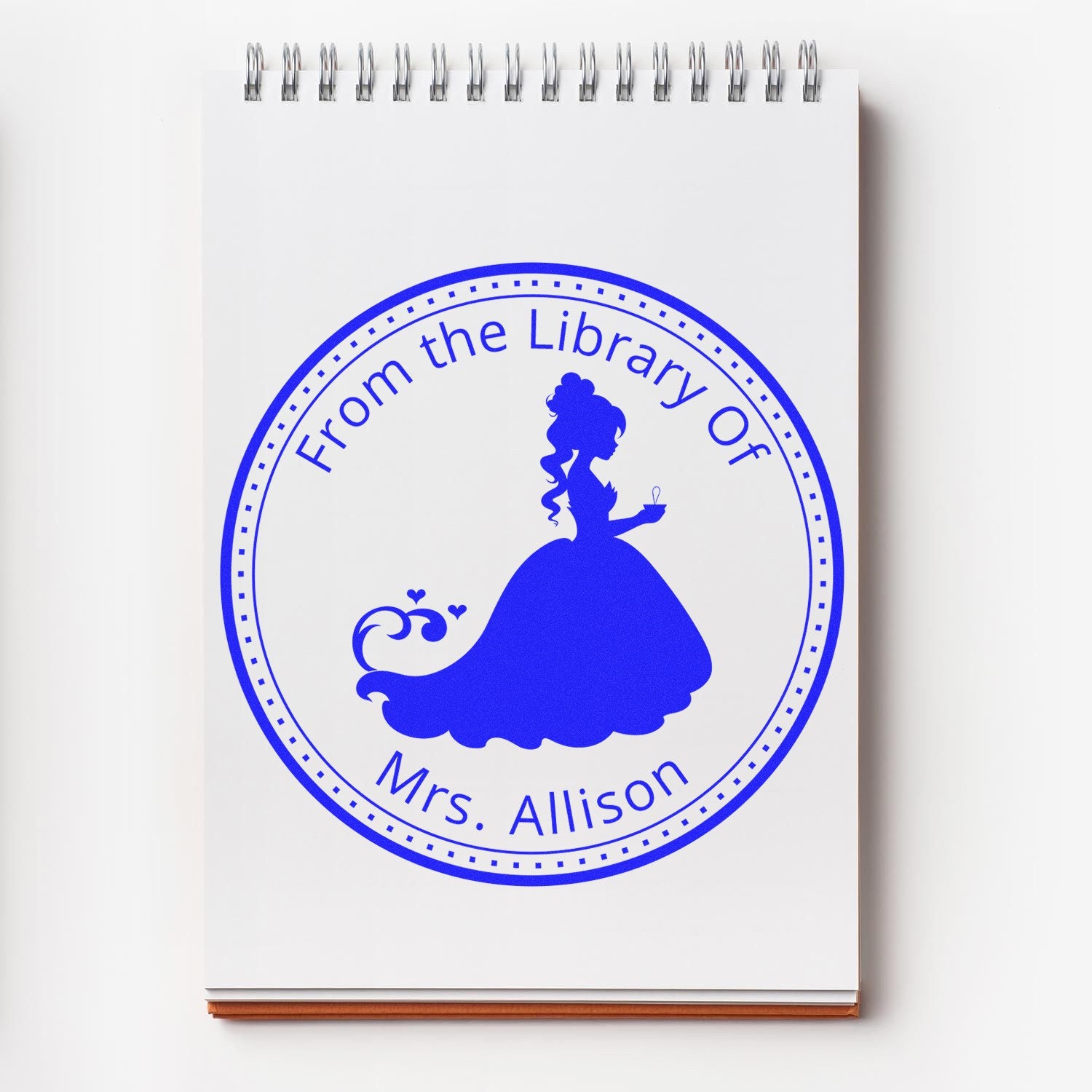 Blue circular library stamp seal on a white spiral notebook, with a woman silhouette and From the Library Of Mrs. Allison.