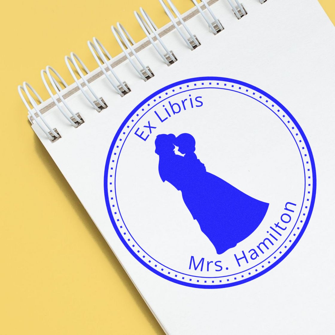 Blue ex libris stamp seal with Mrs. Hamilton silhouette on a white spiral notebook against a yellow background.