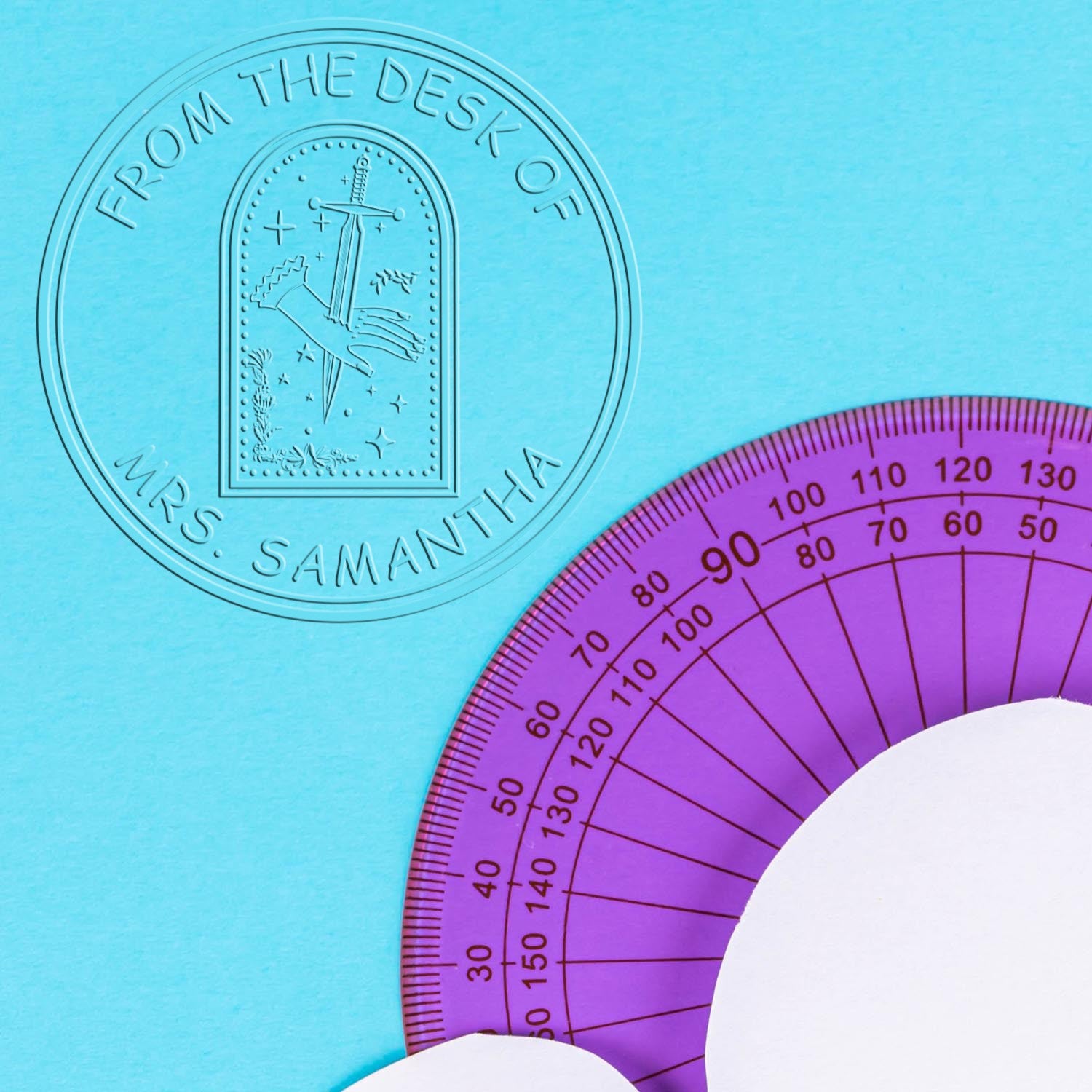 Embossed circular desk seal stamp reading From the Desk of Mrs. Samantha on aqua, with a purple protractor.