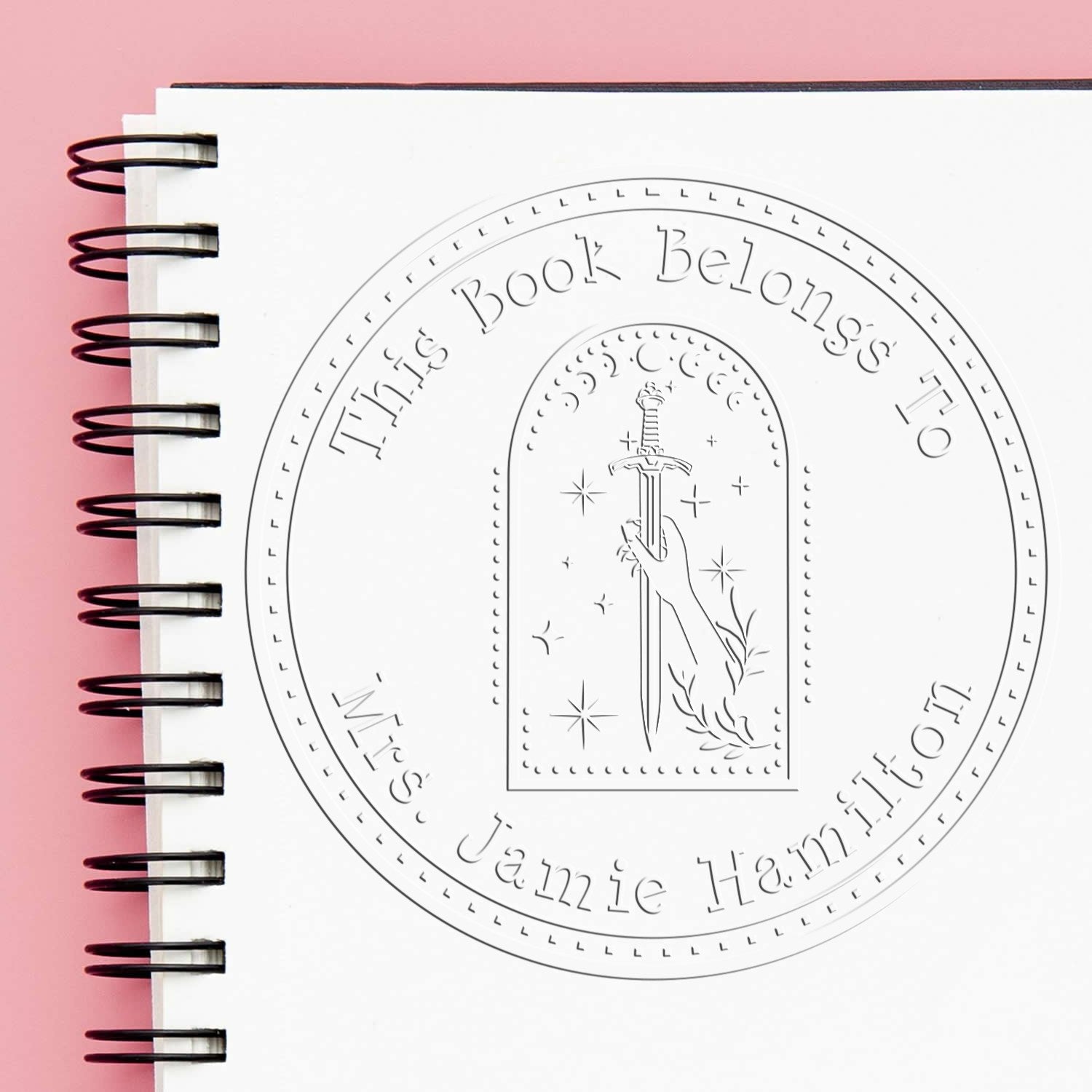 Embossed book seal stamp on white notebook, with circular sword design and This Book Belongs To Mrs. Jamie Hamilton on pink background.