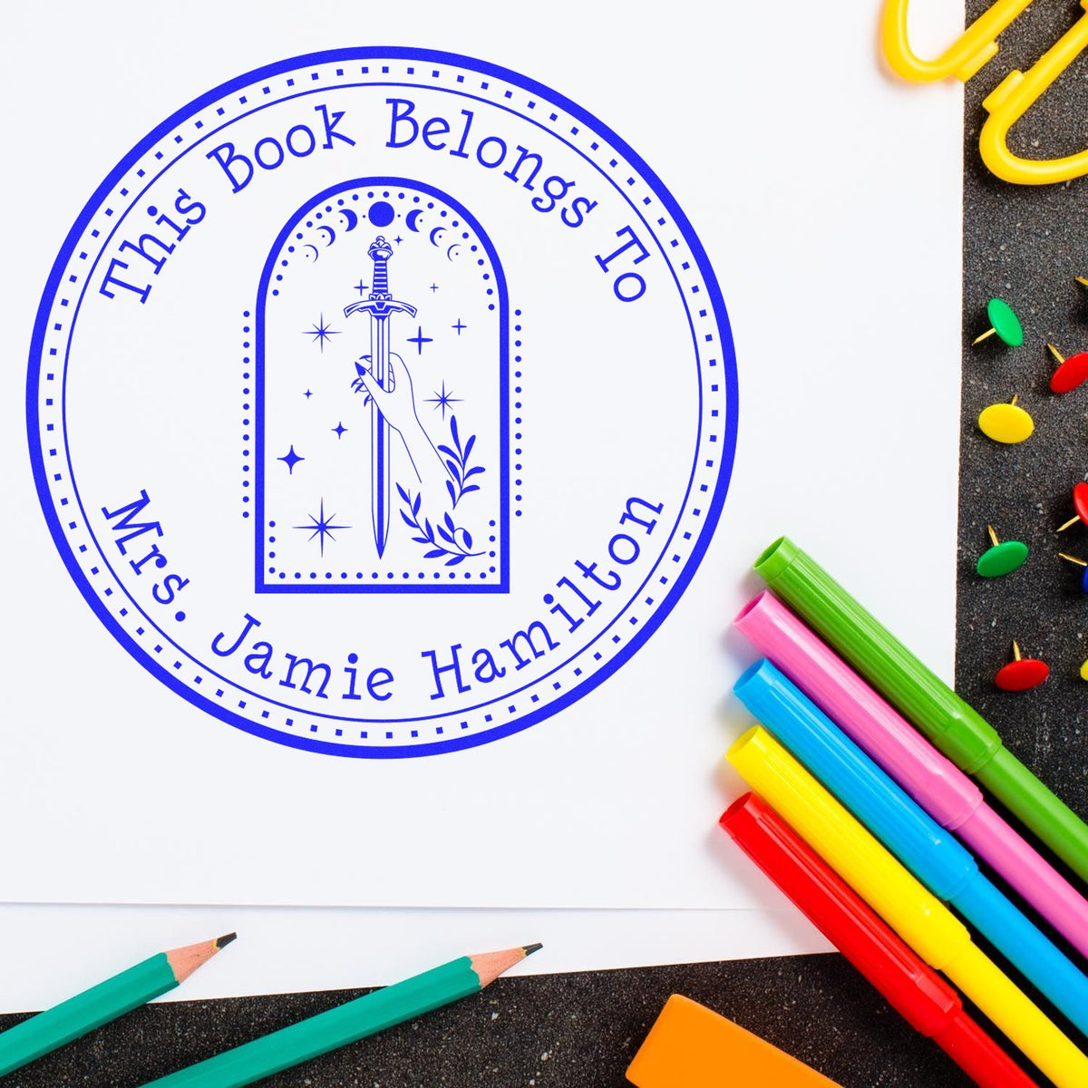 Blue circular book stamp and seal reading This Book Belongs to Mrs. Jamie Hamilton, with sword emblem and colorful markers.