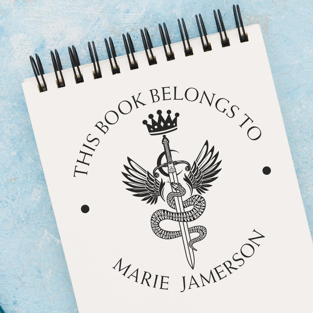 Black custom book-belongs-to stamp seal on white spiral notebook, featuring crown, sword and snake on a blue background.