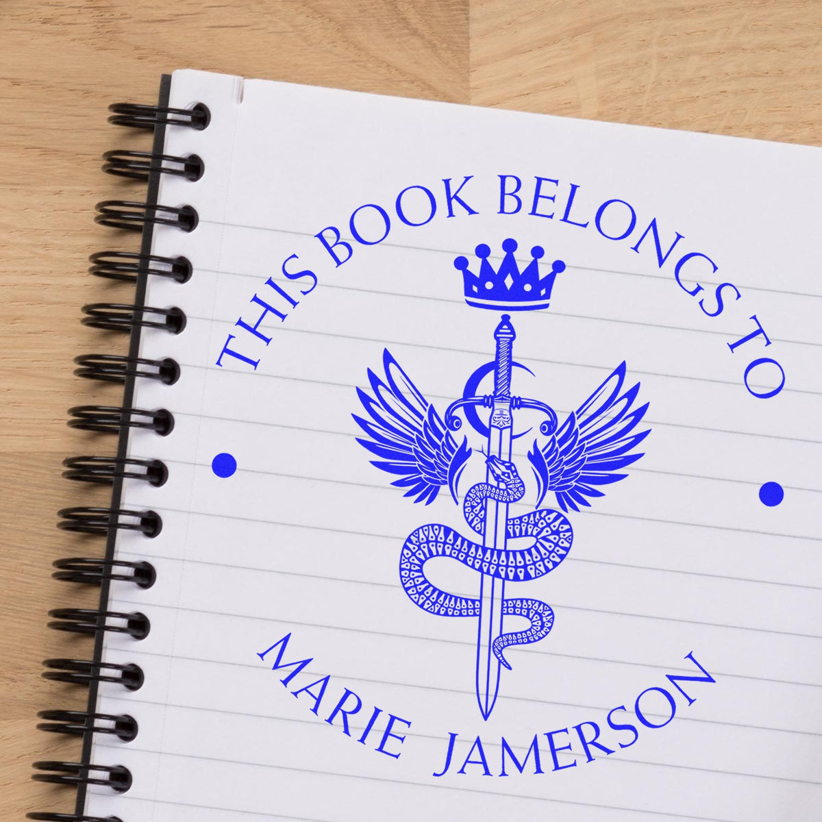 Blue custom book stamp seal with crown, wings and sword on a white spiral notebook over a wood table.