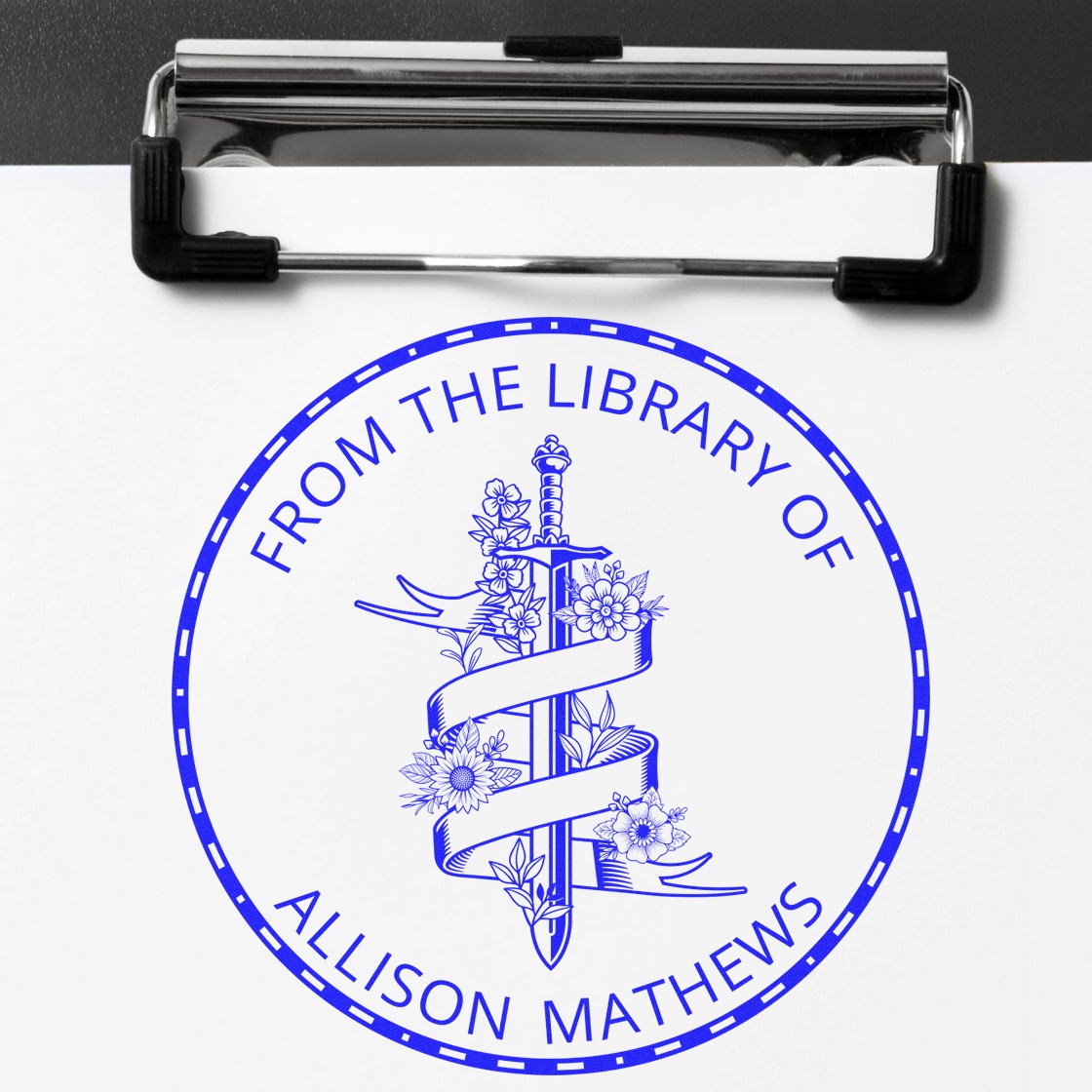 Blue custom library stamp seal with sword and floral crest, centered on white paper clipped to a metal clipboard.