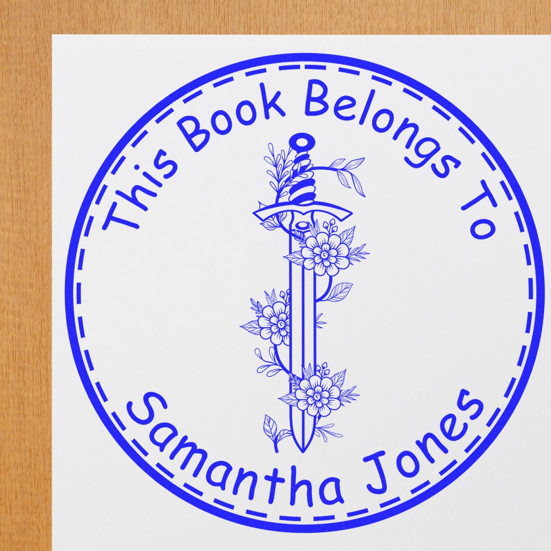 Blue circular personalized book stamp seal reading This Book Belongs To Samantha Jones with floral sword design on white paper.