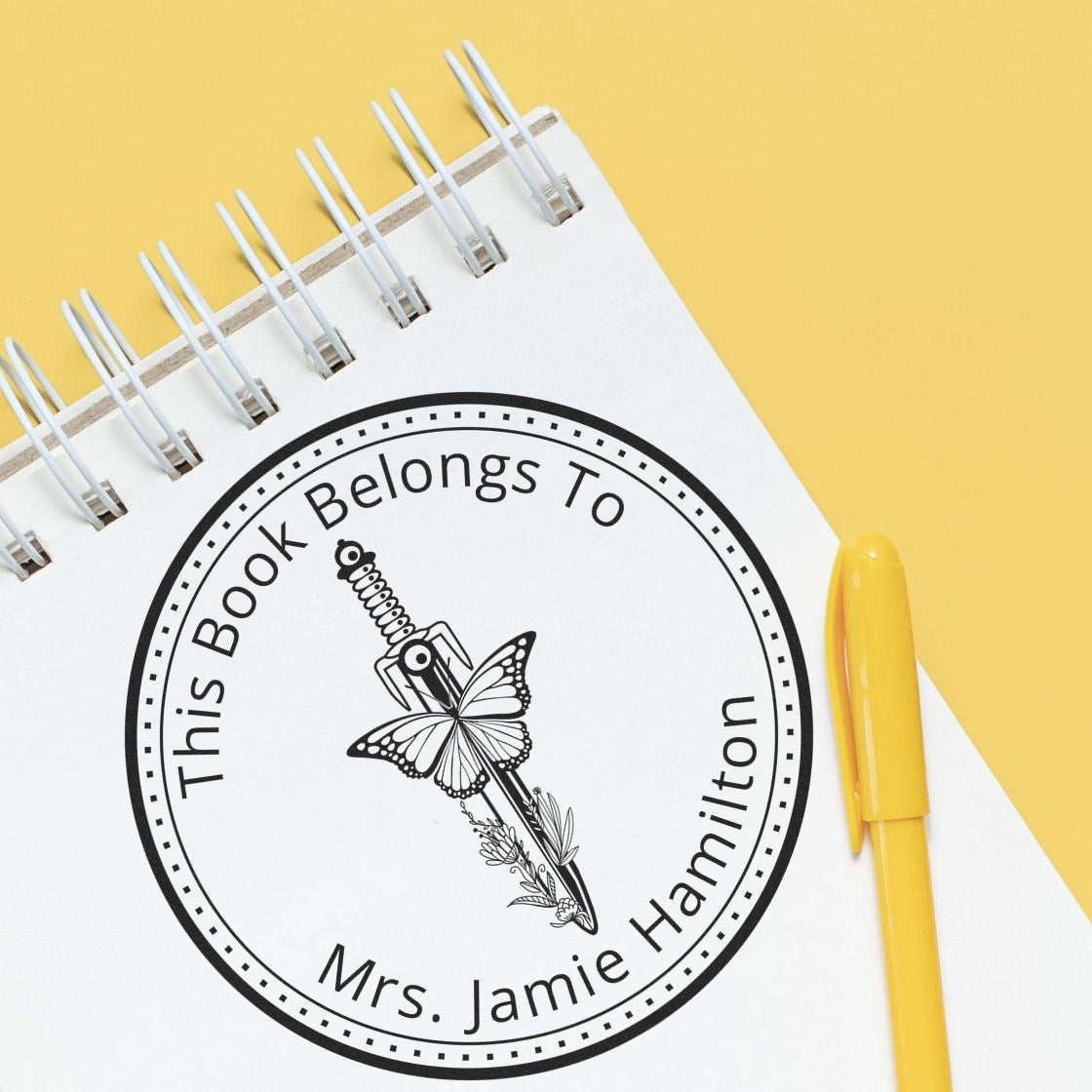 Black personalized stamp/seal for books on white spiral notebook, butterfly sword design, beside yellow pen on yellow background.
